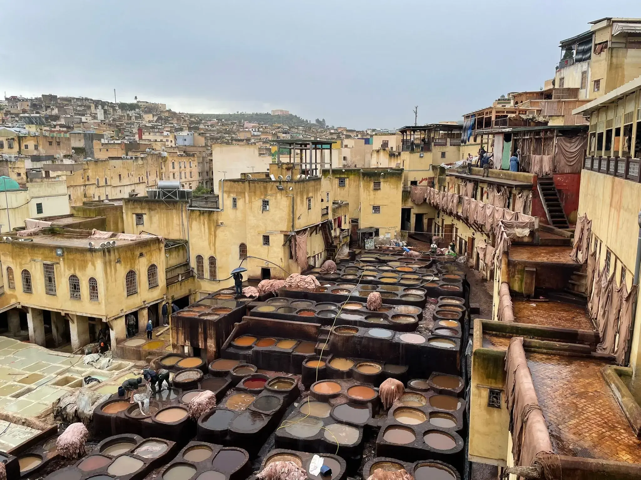 Tannery vats at Chouara Tannery in Fes, Morocco with workers handling hides among dyeing pools under an overcast sky.