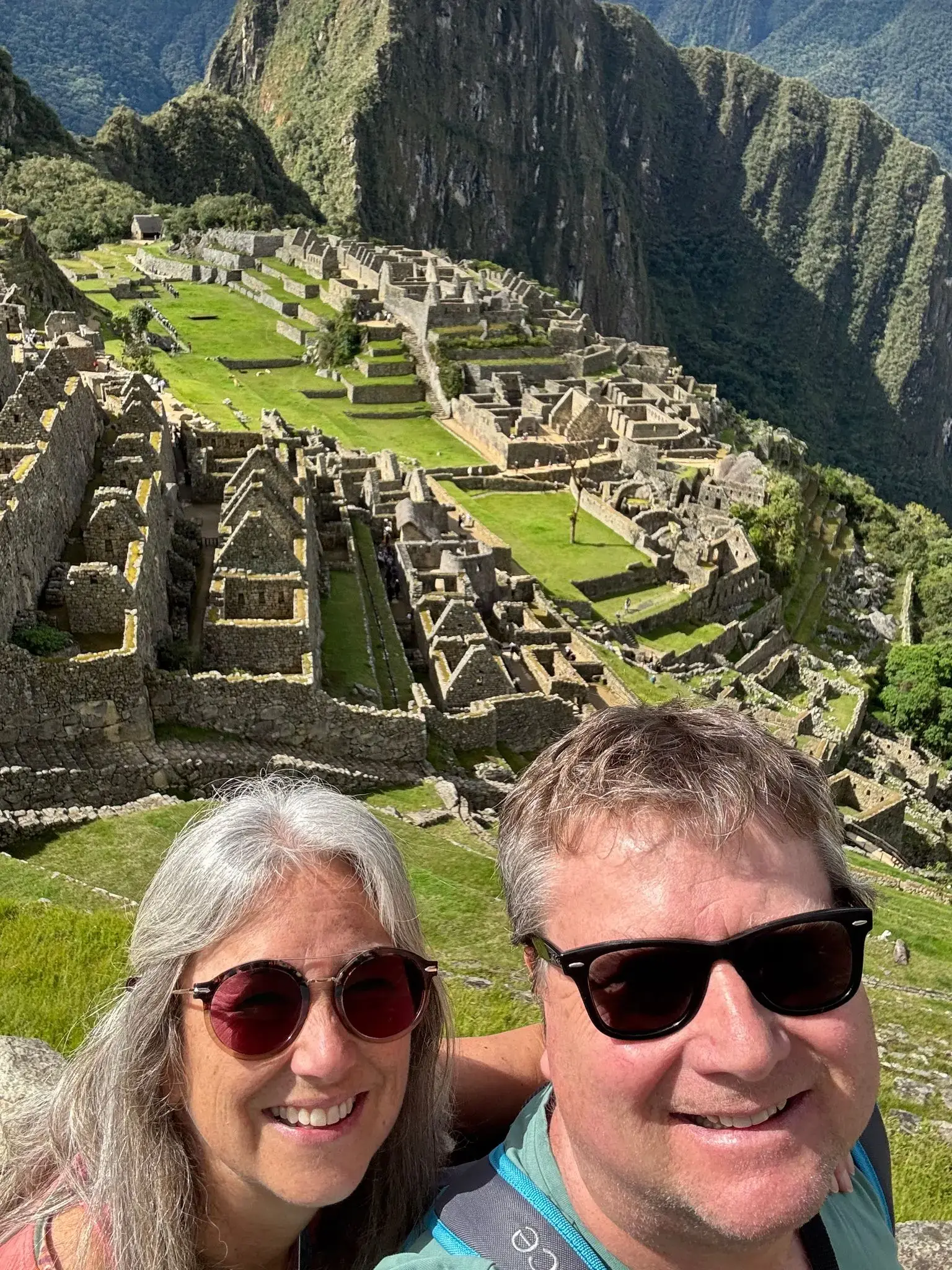 Machu Picchu ruins with two travelers taking a selfie in the foreground, terraces and steep mountains visible, Peru