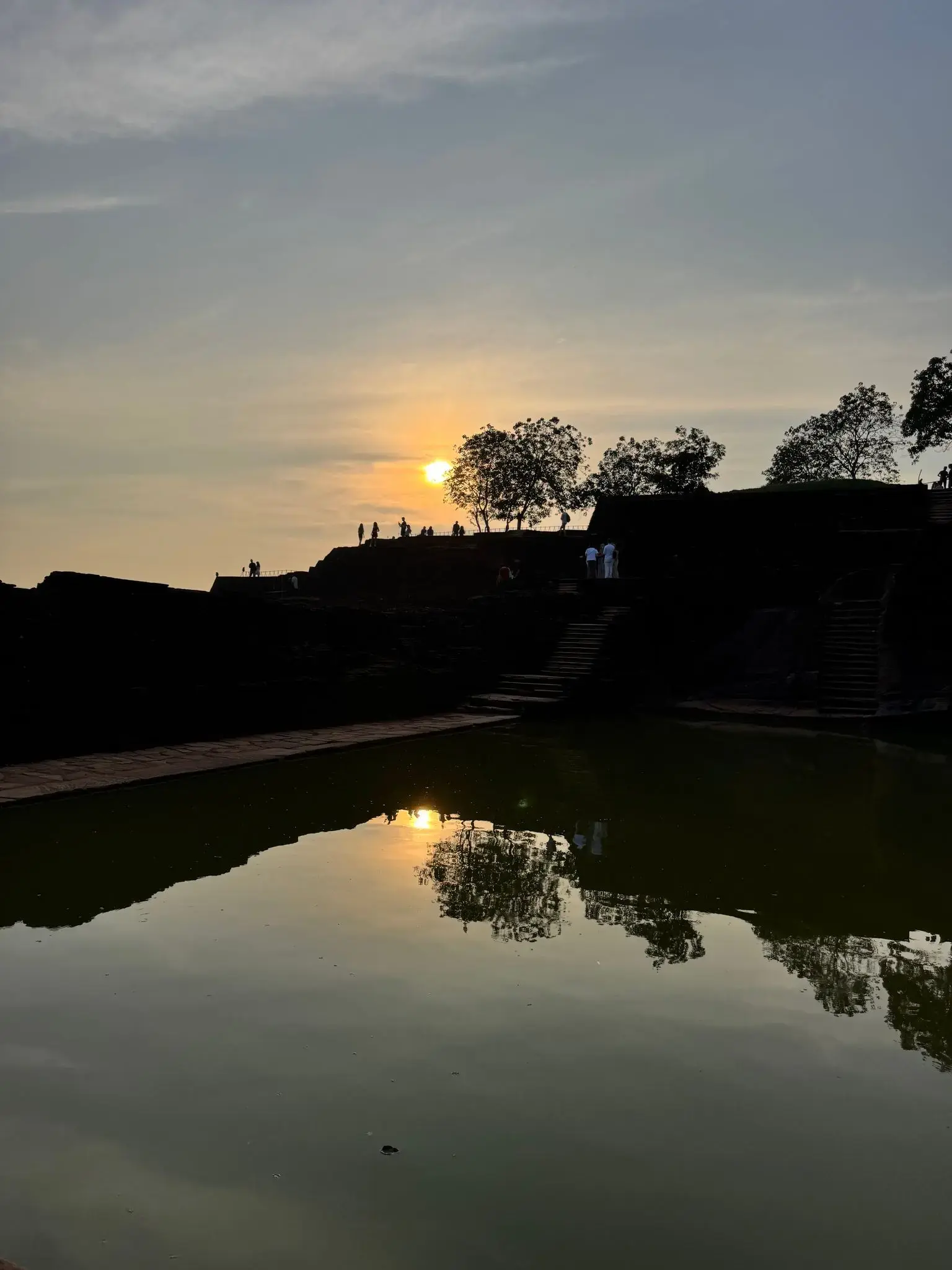 Sigiriya Rock (Lion Rock) silhouetted at sunset with visitors on the summit and its reflecting pool, Sri Lanka.