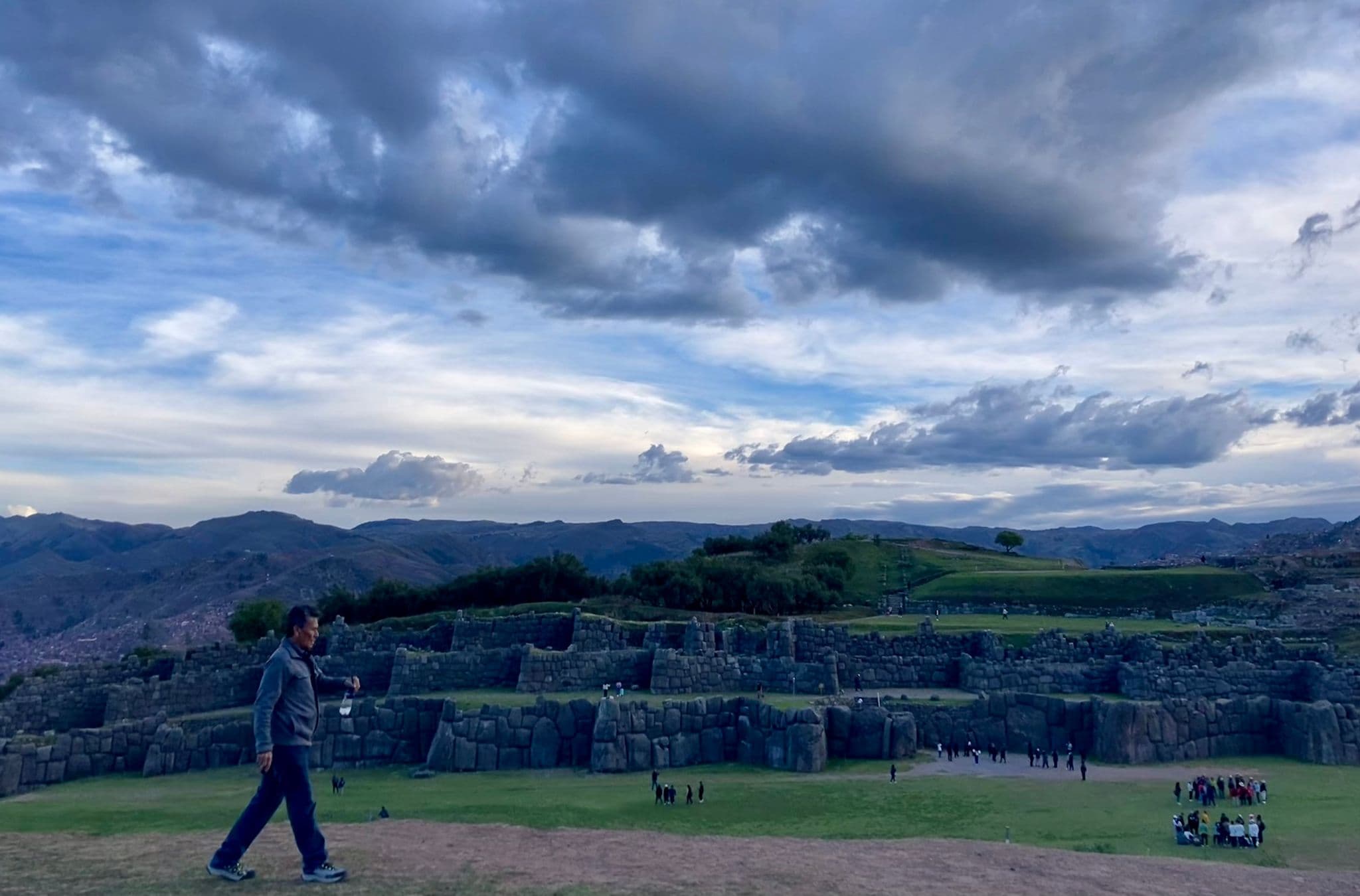 Sacsayhuamán stone terraces and walls with a man walking in the foreground, mountains and cloudy sky near Cusco, Peru.