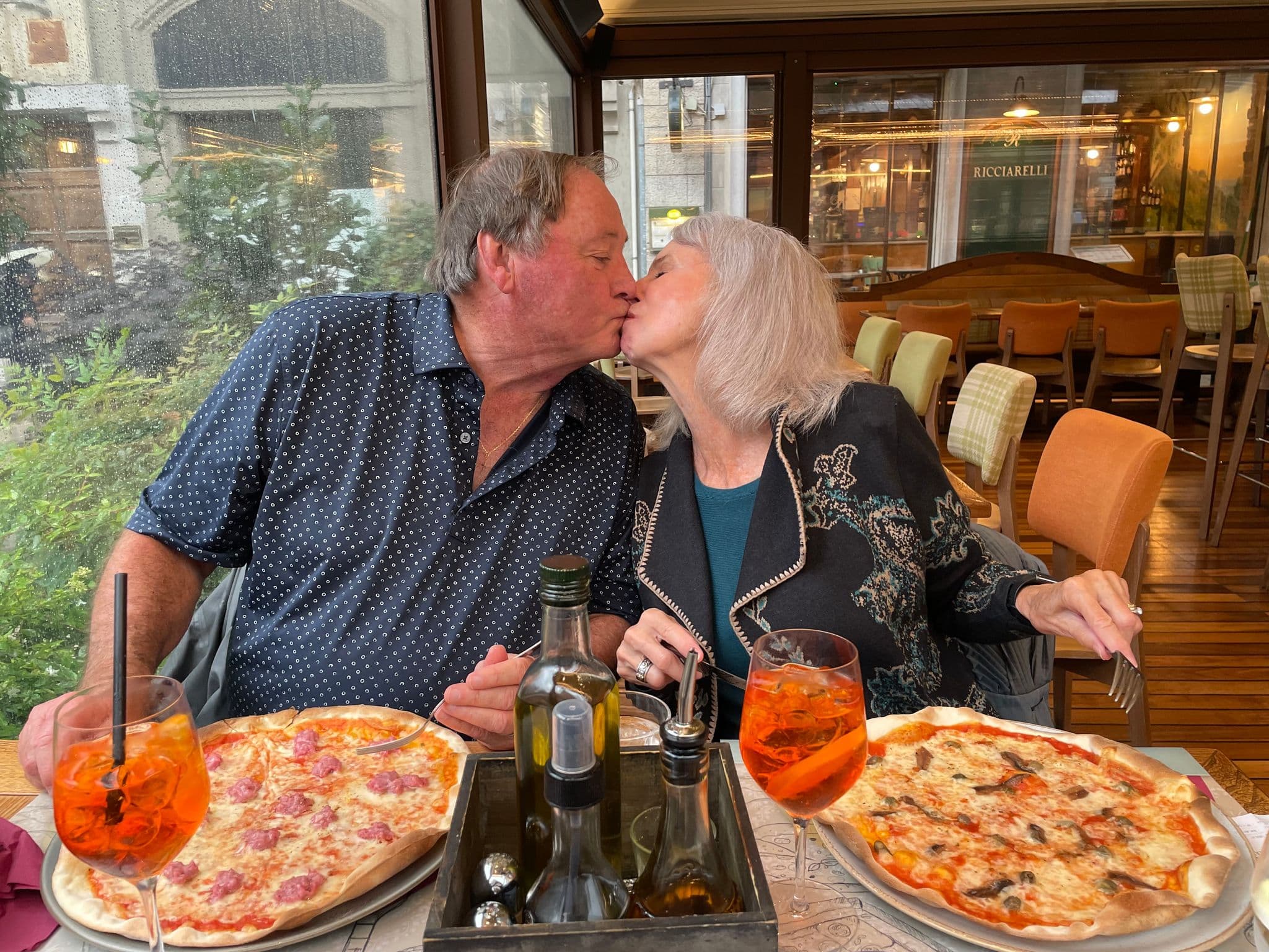 An older couple kissing across a table with two pizzas and Aperol spritz drinks in a restaurant in Montecatini Terme, Italy.