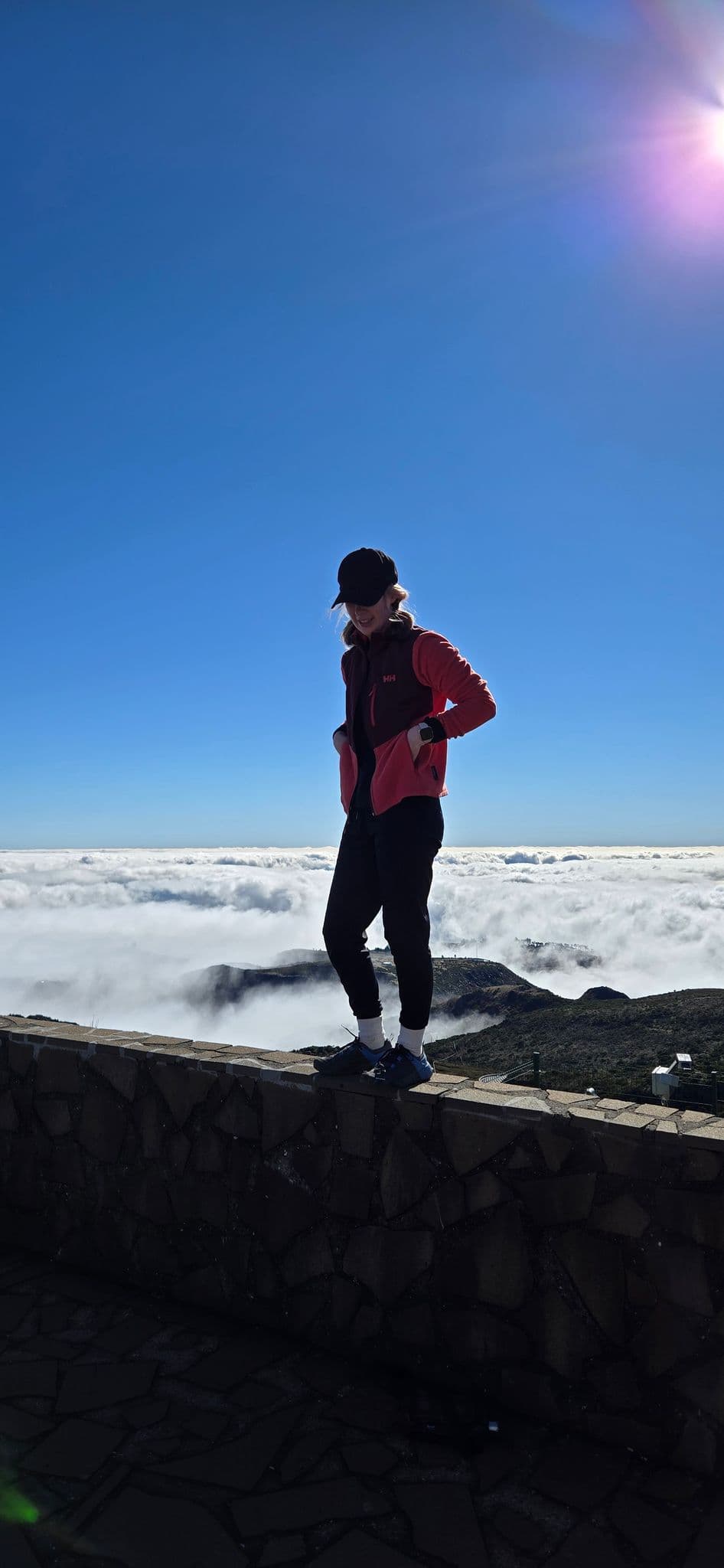 Person standing on a stone lookout wall above a sea of clouds with mountain ridges below and a clear blue sky.