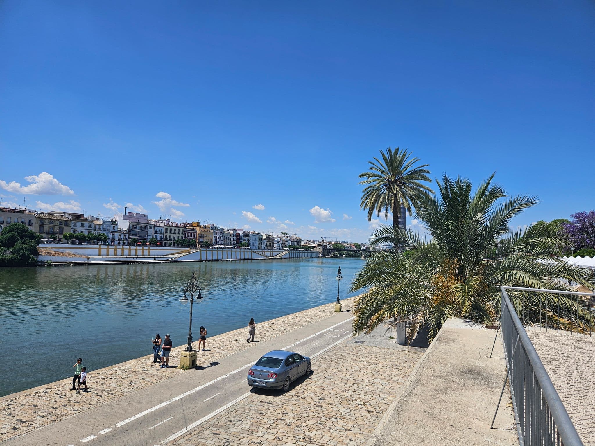 Guadalquivir River flanked by palm trees and a riverside promenade with pedestrians in Seville, Spain.