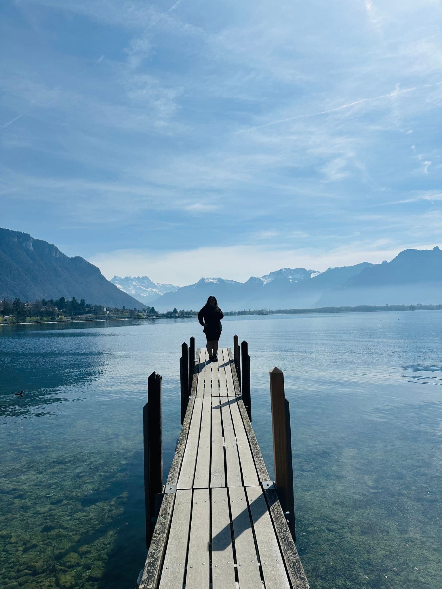 Wooden pier leading to a person standing at the end with the Alps over a clear lake, Switzerland.