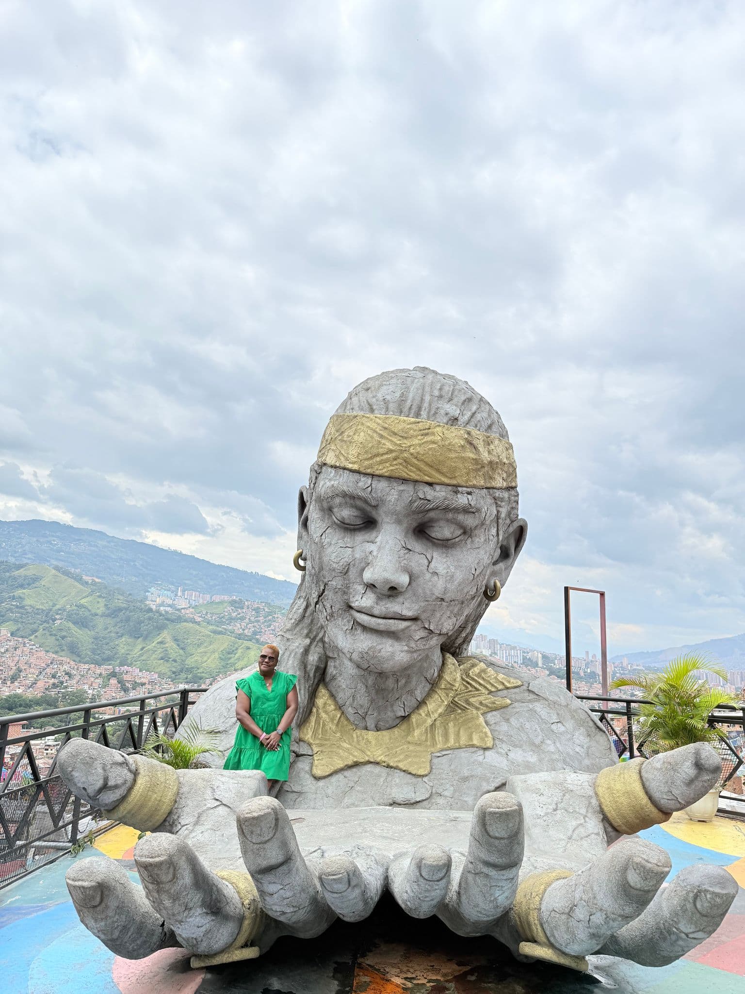 Giant stone sculpture with outstretched hands and a person leaning on its arm, overlooking Medellín, Colombia