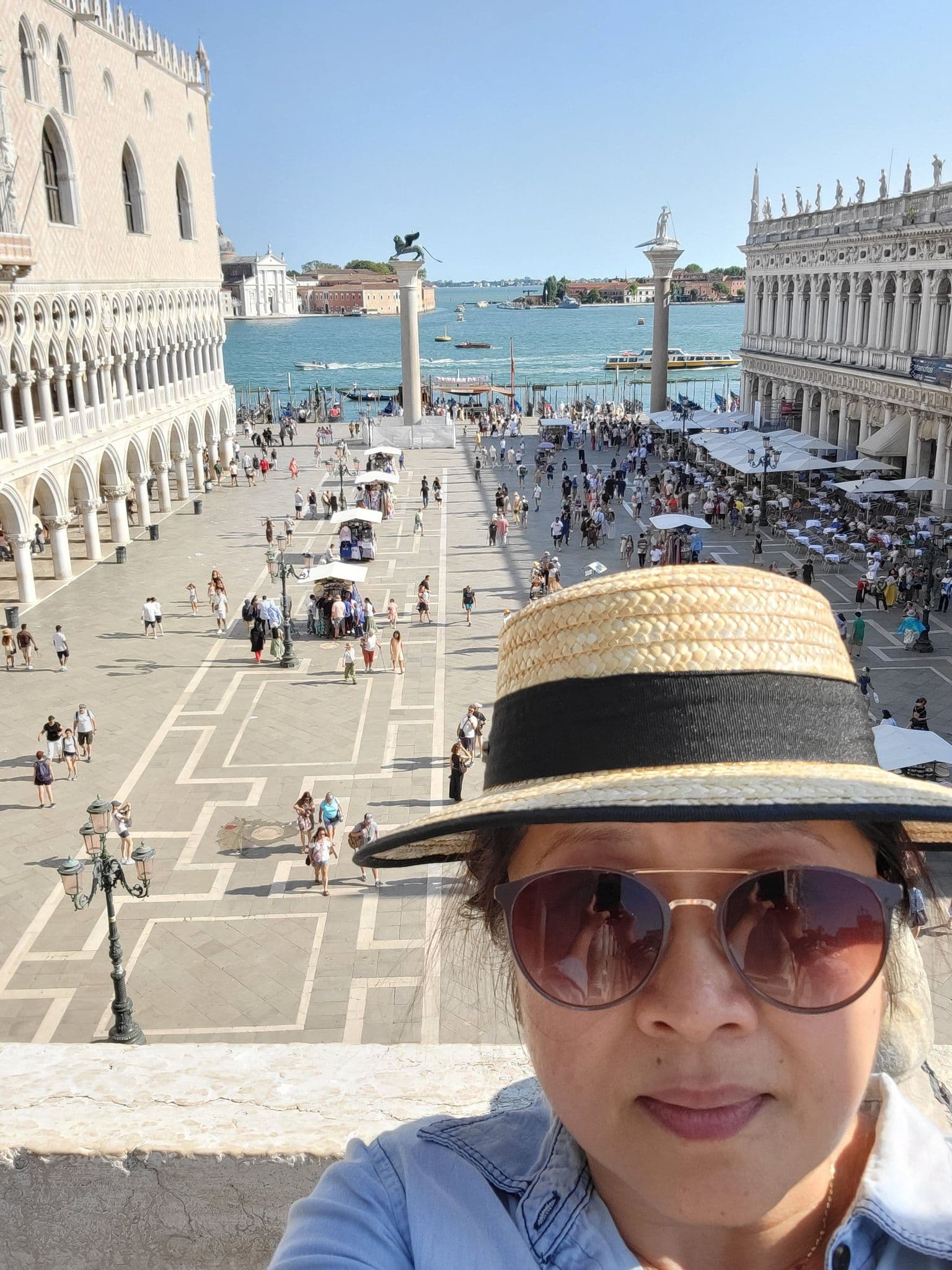 View of Piazza San Marco from St. Mark's Basilica rooftop in Venice, Italy, with a traveler selfie wearing a straw hat and sunglasses.