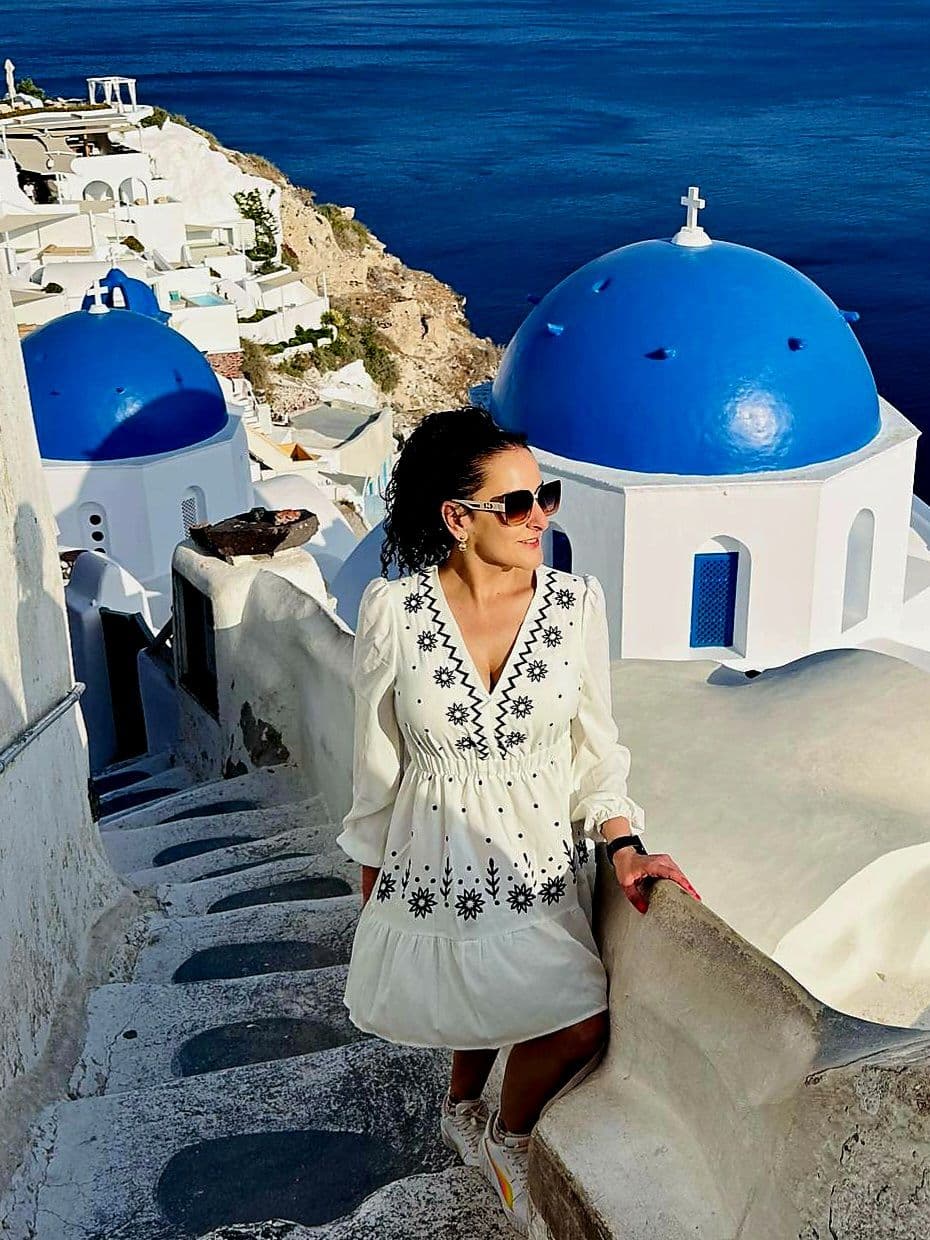 Blue-domed churches in Oia, Santorini with a woman in a white dress standing on stone steps overlooking the Aegean Sea, Greece.