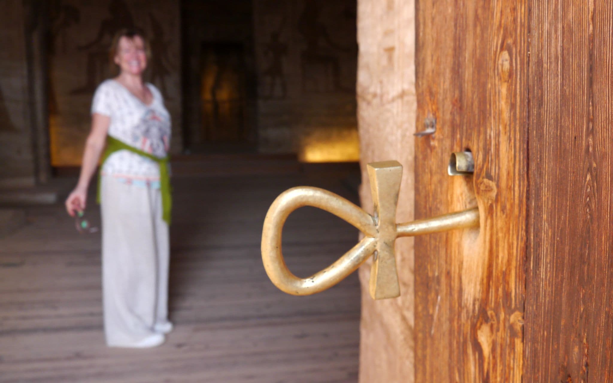 Ankh-shaped brass door handle at Abu Simbel, Egypt, with a blurred traveler standing inside the temple.