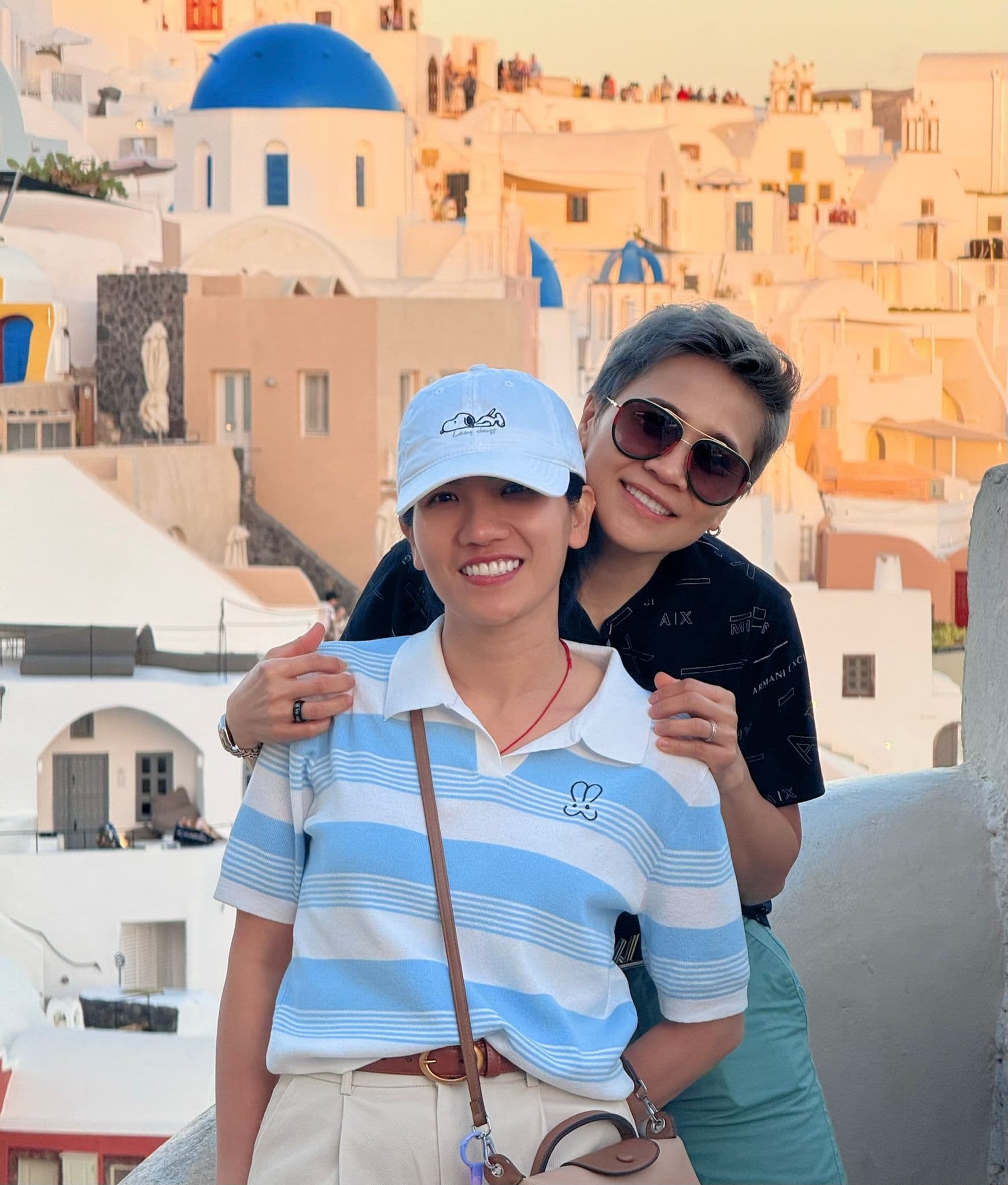 Blue-domed buildings in Oia, Santorini, Greece with two travelers smiling and posing on a terrace.
