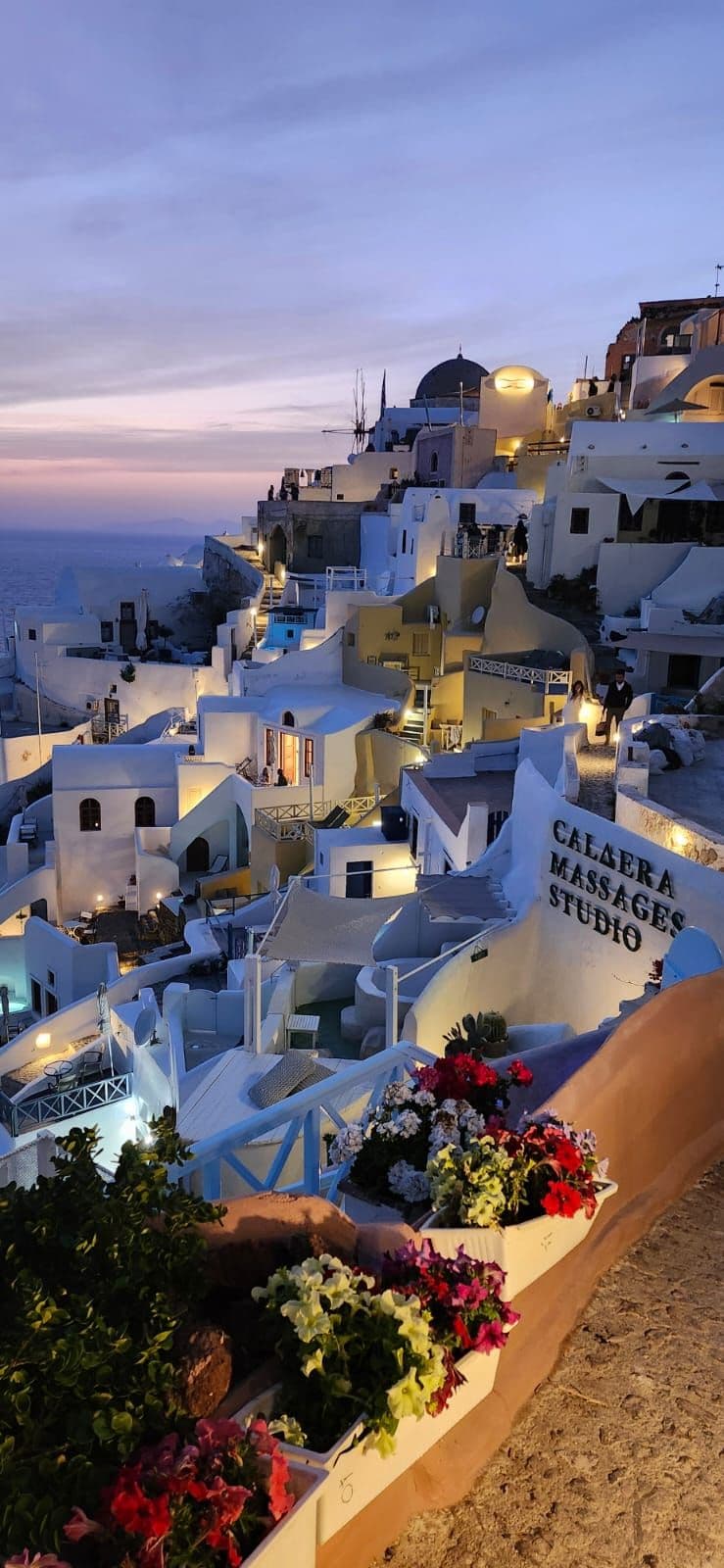Oia's whitewashed cliffside buildings at sunset overlooking the caldera, with lit terraces and flower boxes along a walkway, Santorini, Greece.