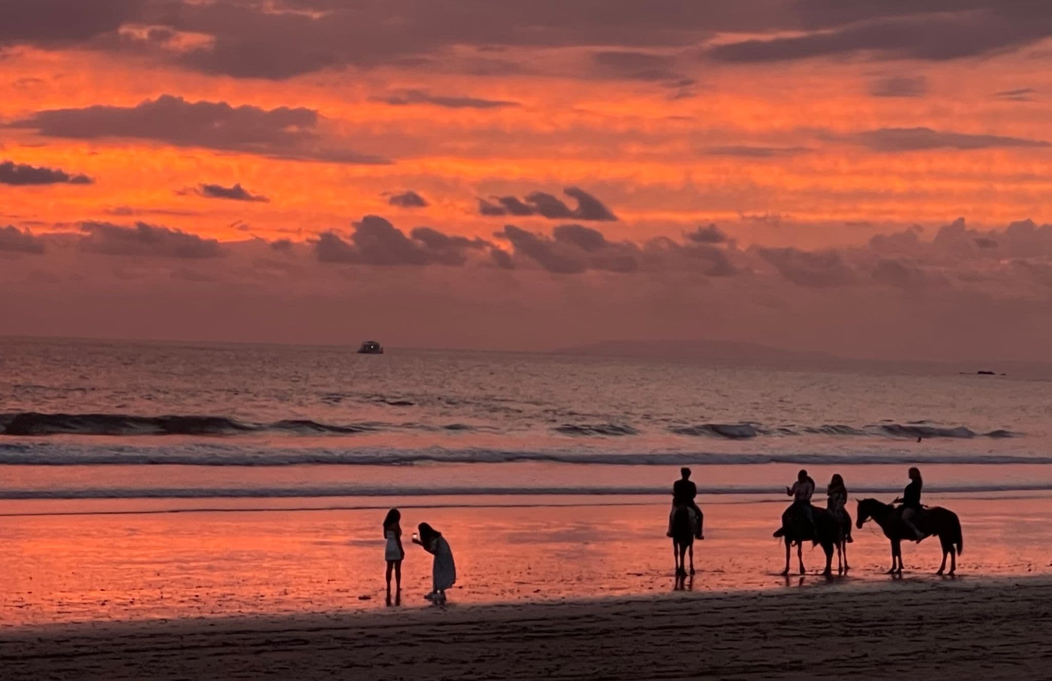 Sunset over Jacó Beach, Costa Rica, with silhouetted people and horseback riders reflected on wet sand.