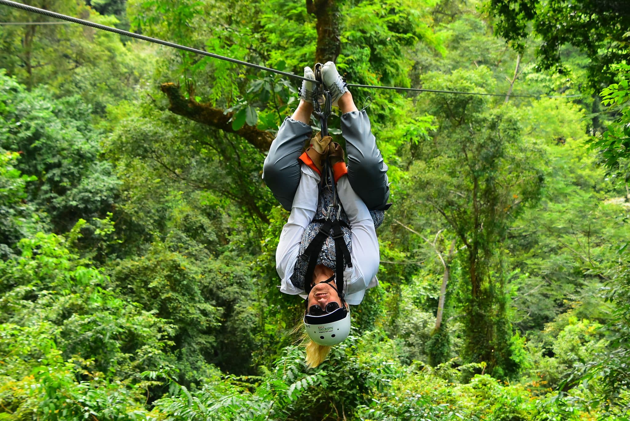 Person wearing a helmet and sunglasses ziplining upside-down through a dense rainforest canopy in Costa Rica.