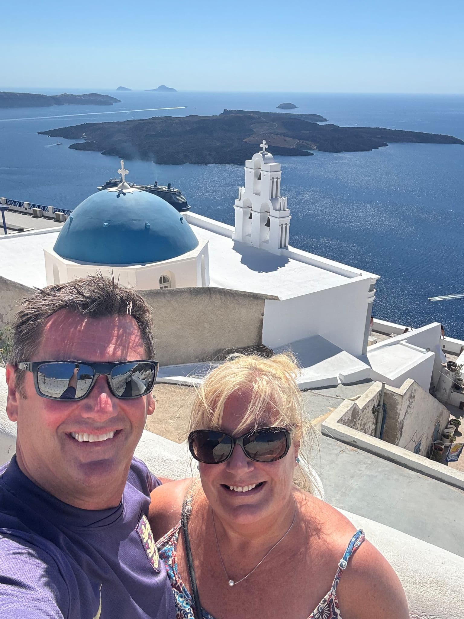 Blue-domed church and bell tower overlooking the caldera in Oia, Santorini, Greece, with two travelers posing for a selfie.