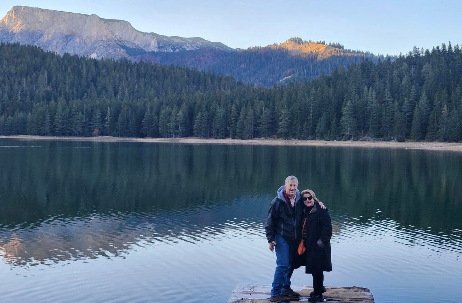 Black Lake (Crno Jezero) with two travelers standing on a wooden dock at the water's edge near Žabljak, Montenegro.
