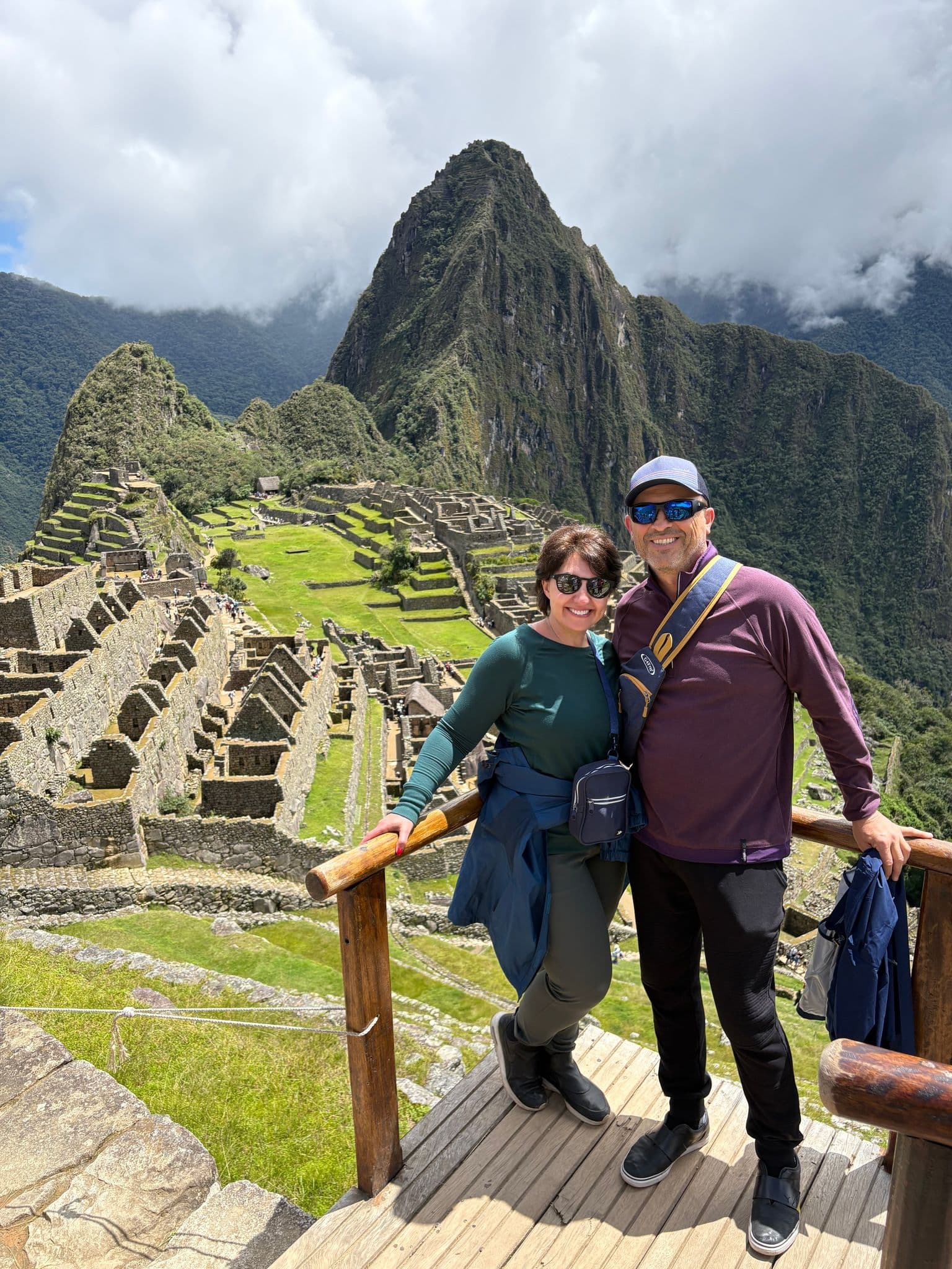 Machu Picchu ruins and Huayna Picchu peak in the Cusco region, Peru, with two travelers posing on a wooden overlook.