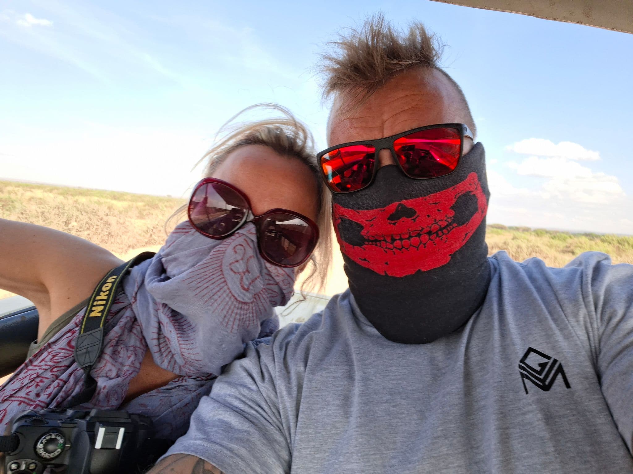 Two people wearing bandanas and sunglasses taking a selfie in an open jeep across dry grassland on a trip.