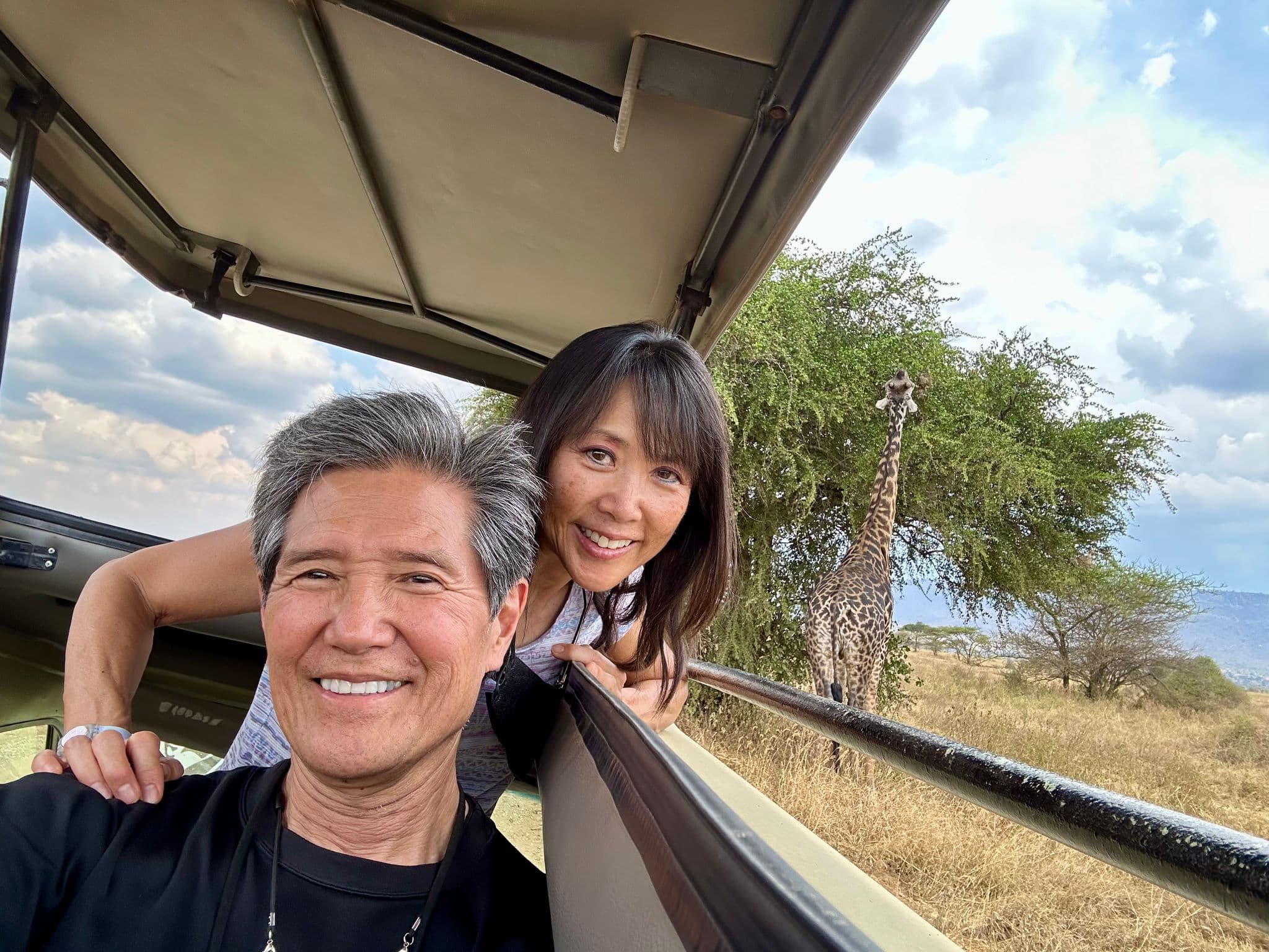 Giraffe standing beside an open-top safari jeep while two travelers lean out smiling during a game drive in Kenya.