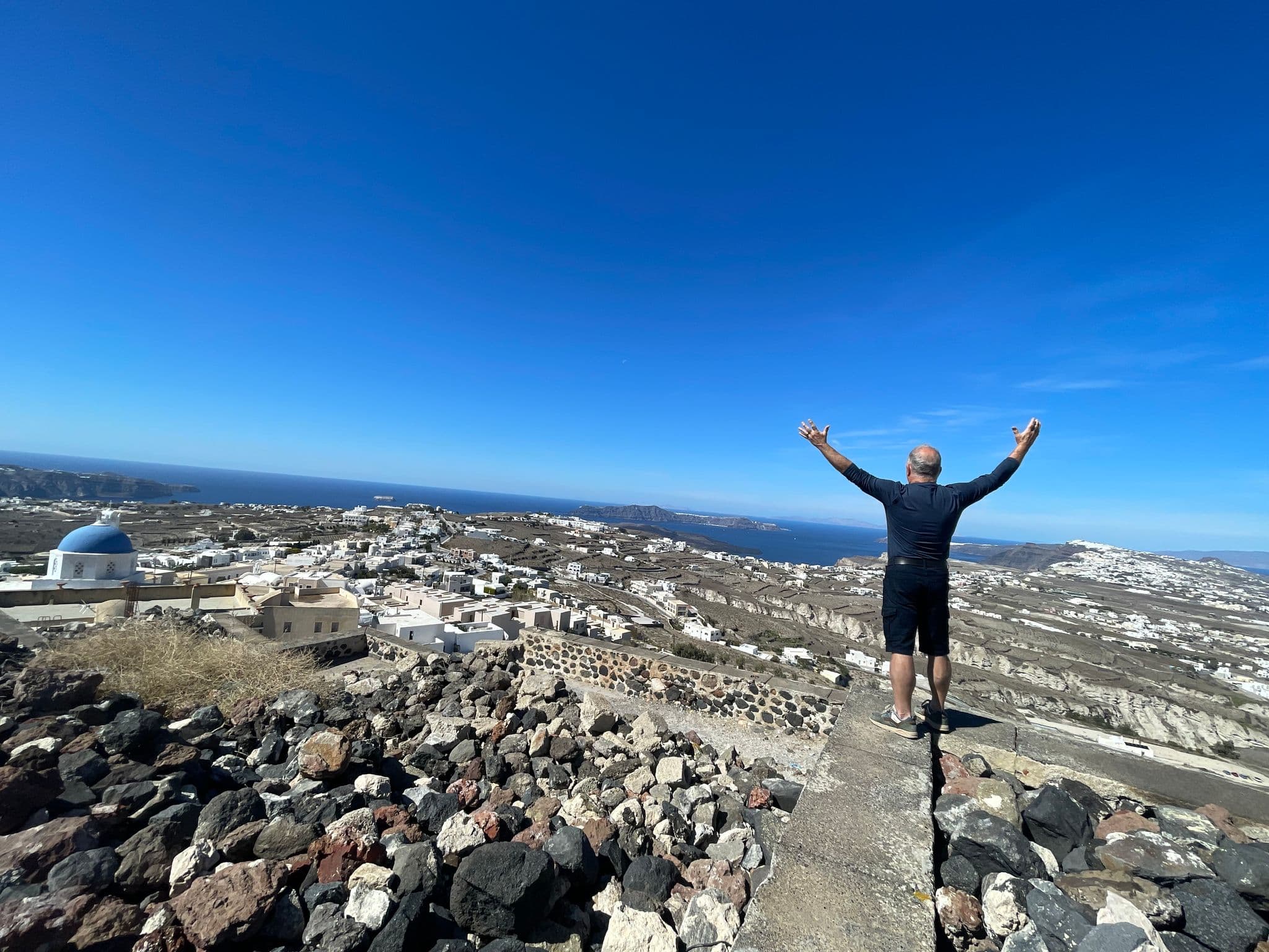 Blue-domed church and Santorini clifftop with a traveler raising his arms overlooking the Aegean, Santorini, Greece.