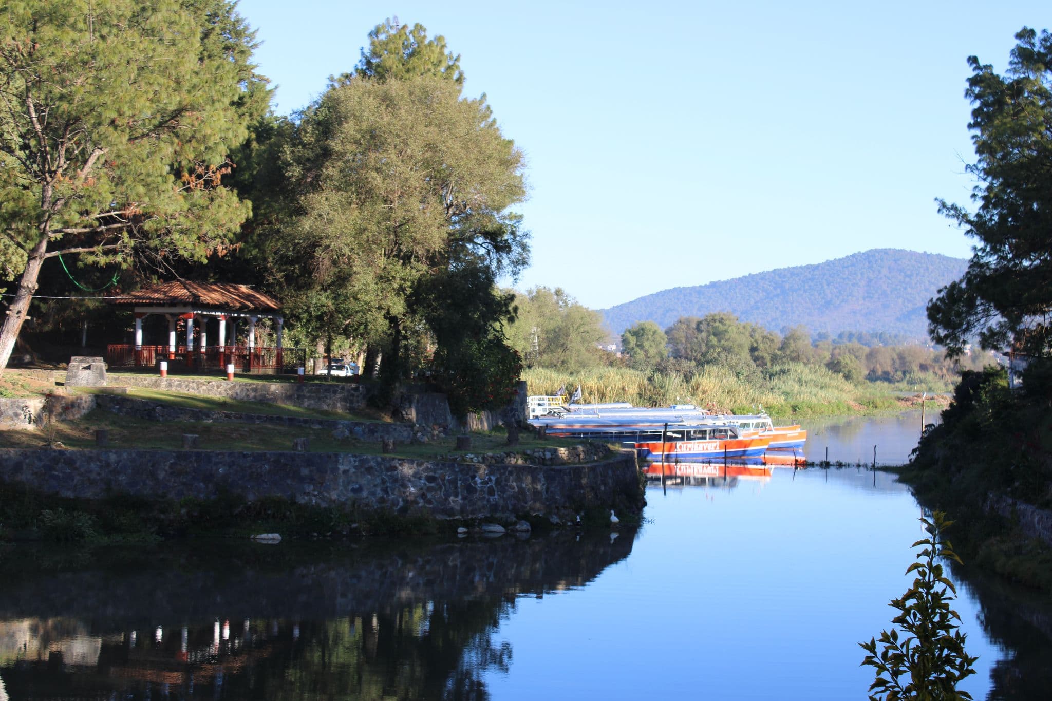 Ferry boats moored along Lake Pátzcuaro with a tree-lined shore and a small pavilion near Isla de Janitzio, Mexico.