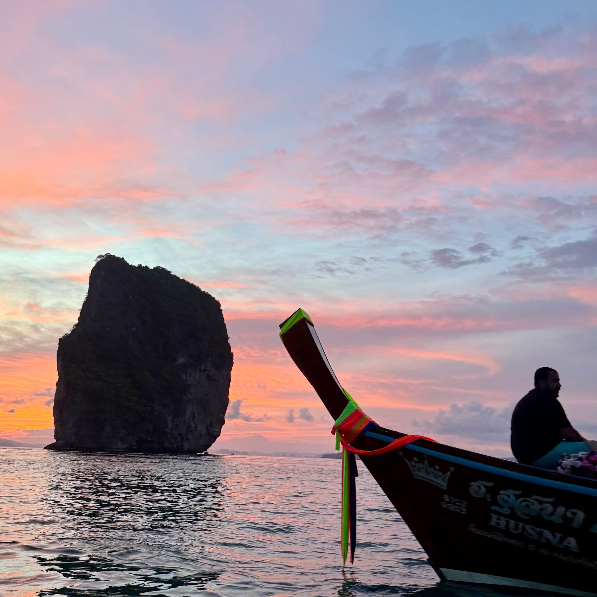 Longtail boat bow with colorful ribbons and a silhouetted passenger beside a towering limestone karst at sunset, Thailand.