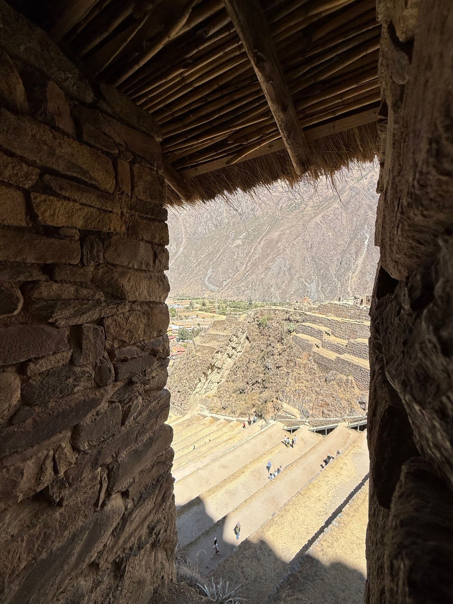 Stone-framed viewpoint looking over terraced slopes of Ollantaytambo in the Sacred Valley, Peru, with small groups of visitors on the paths.