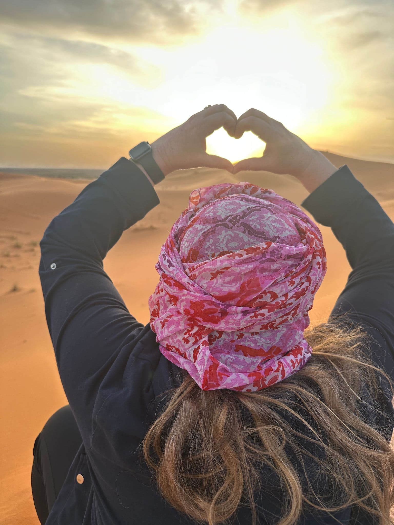 Sahara Desert sand dunes at sunset with a traveler wearing a patterned headscarf forming a heart with their hands.