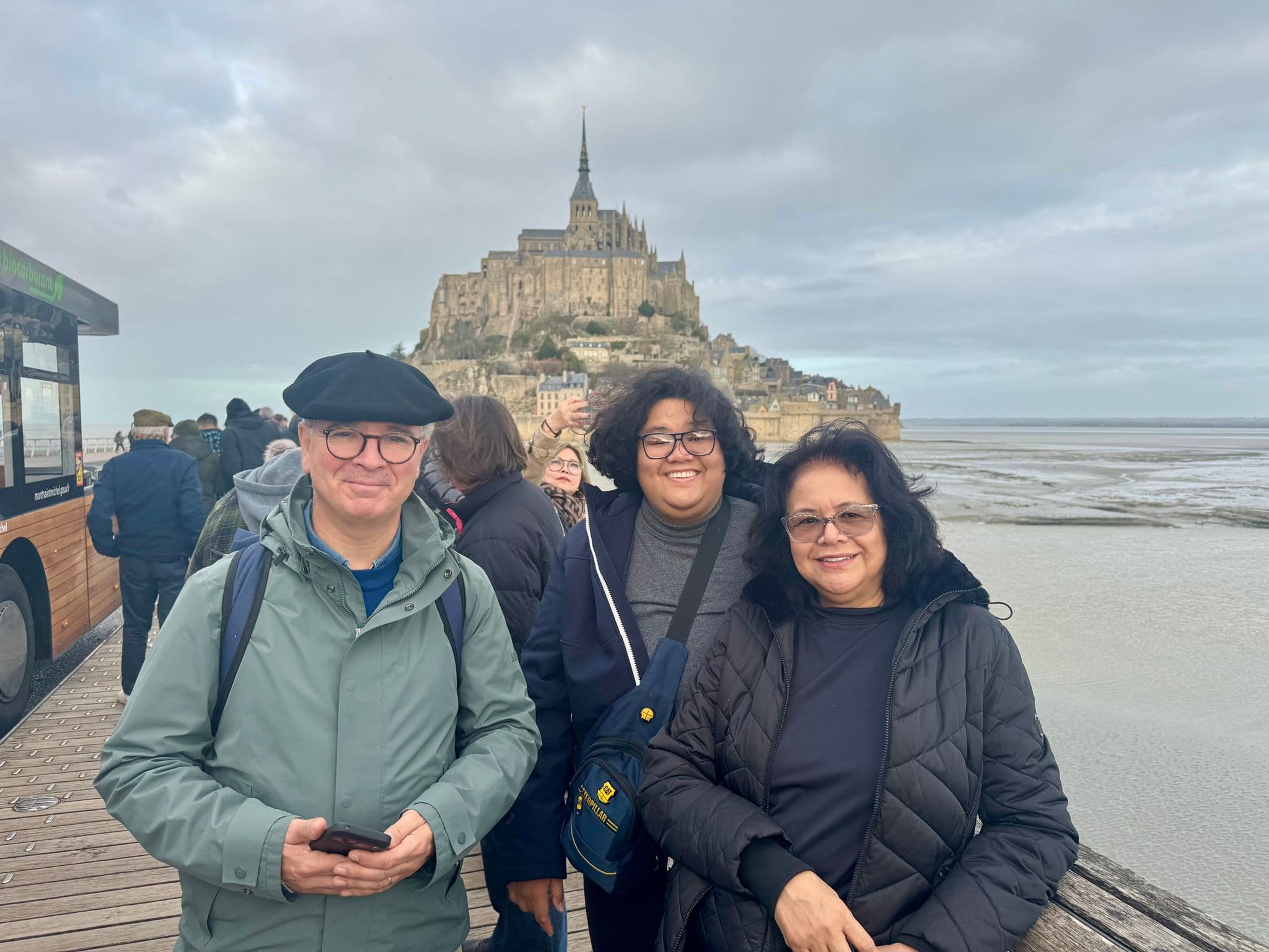 Mont Saint-Michel Abbey rising on its tidal island with three travelers smiling on the wooden causeway in Normandy, France.