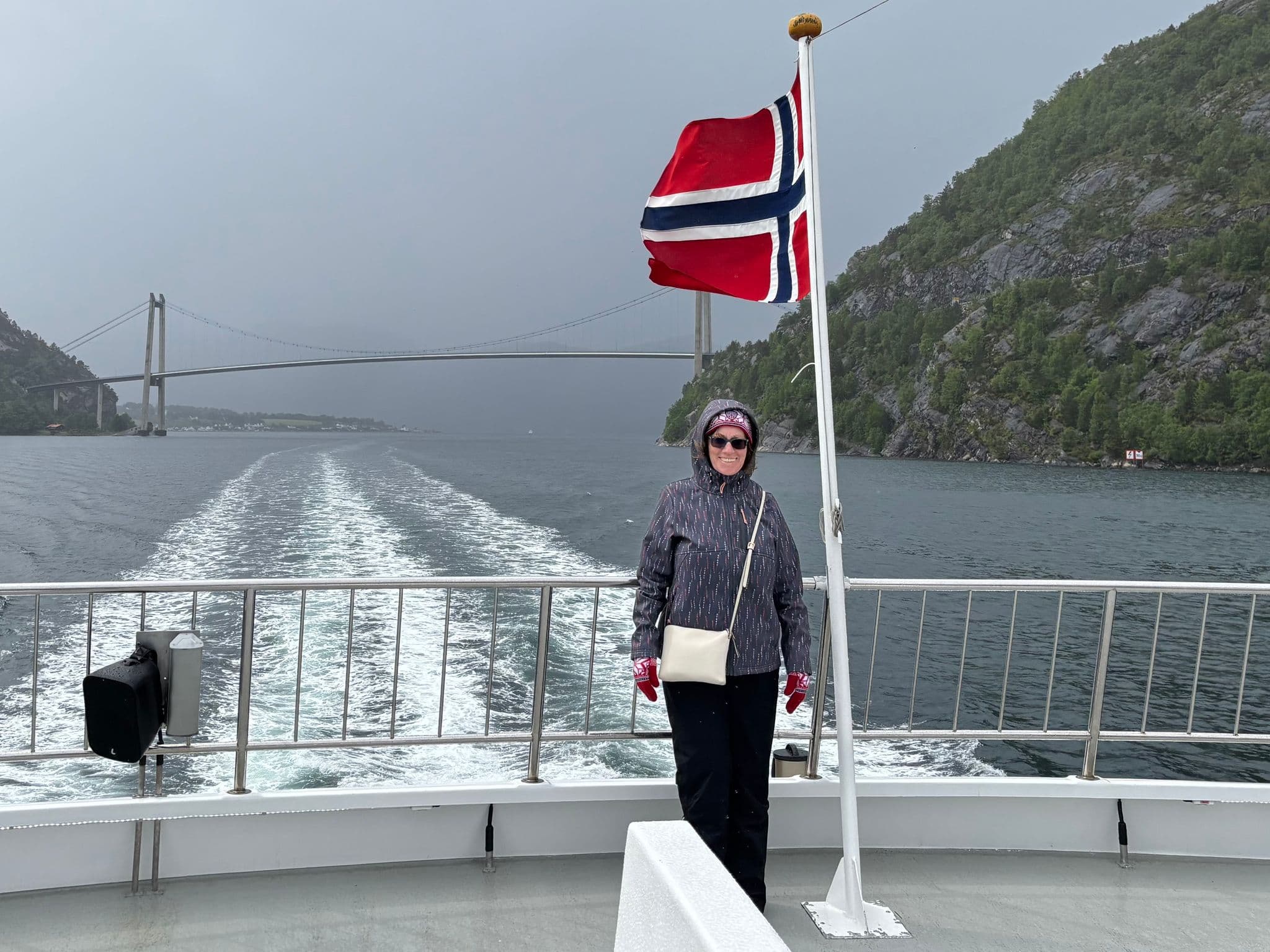 Norwegian flag and a woman standing on a ferry deck with the boat wake, a suspension bridge, and rocky fjord cliffs in Norway.