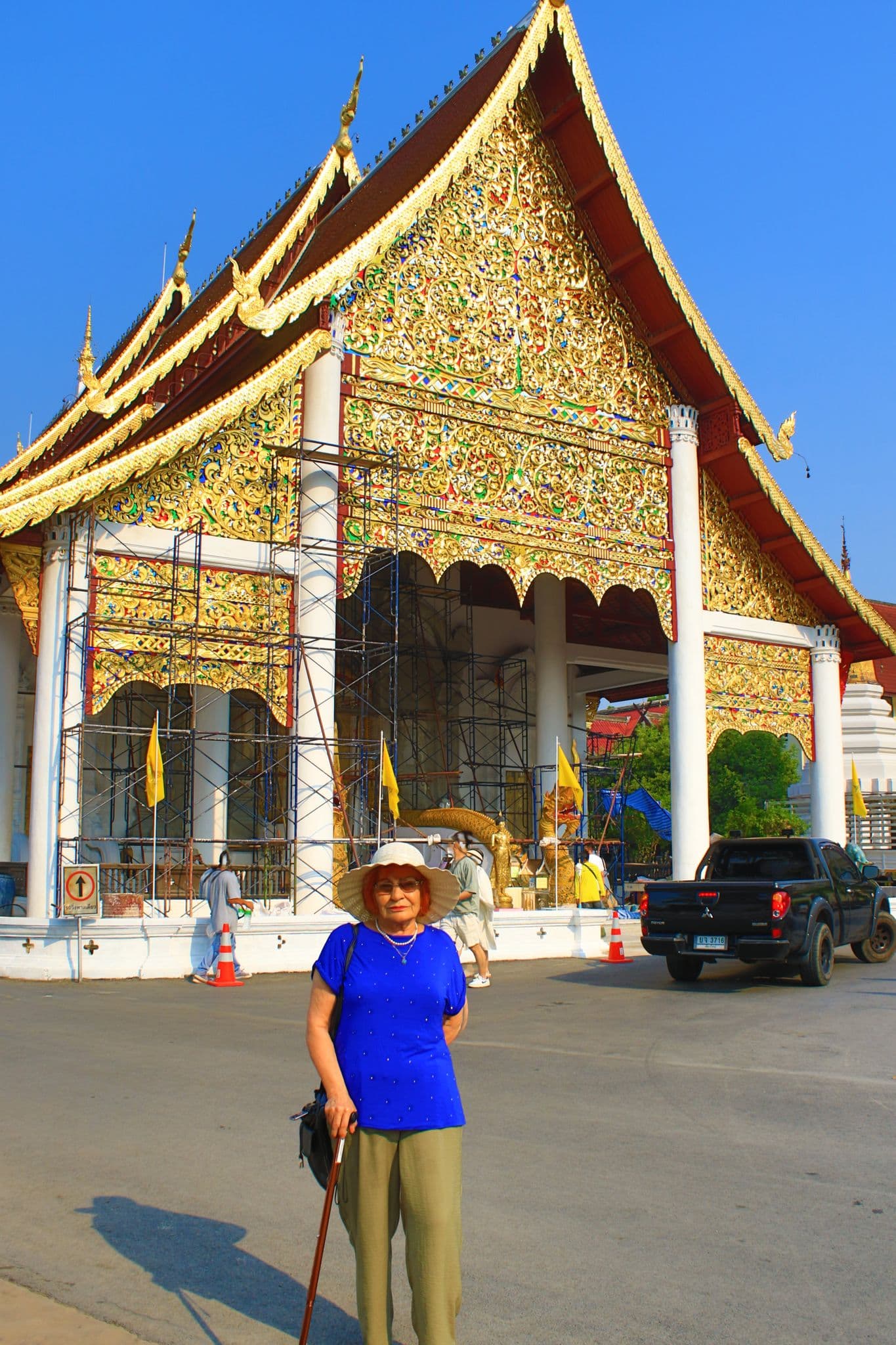 Gilded Thai temple gable with scaffolding and a woman in a blue shirt and sunhat standing with a cane in Bangkok, Thailand.