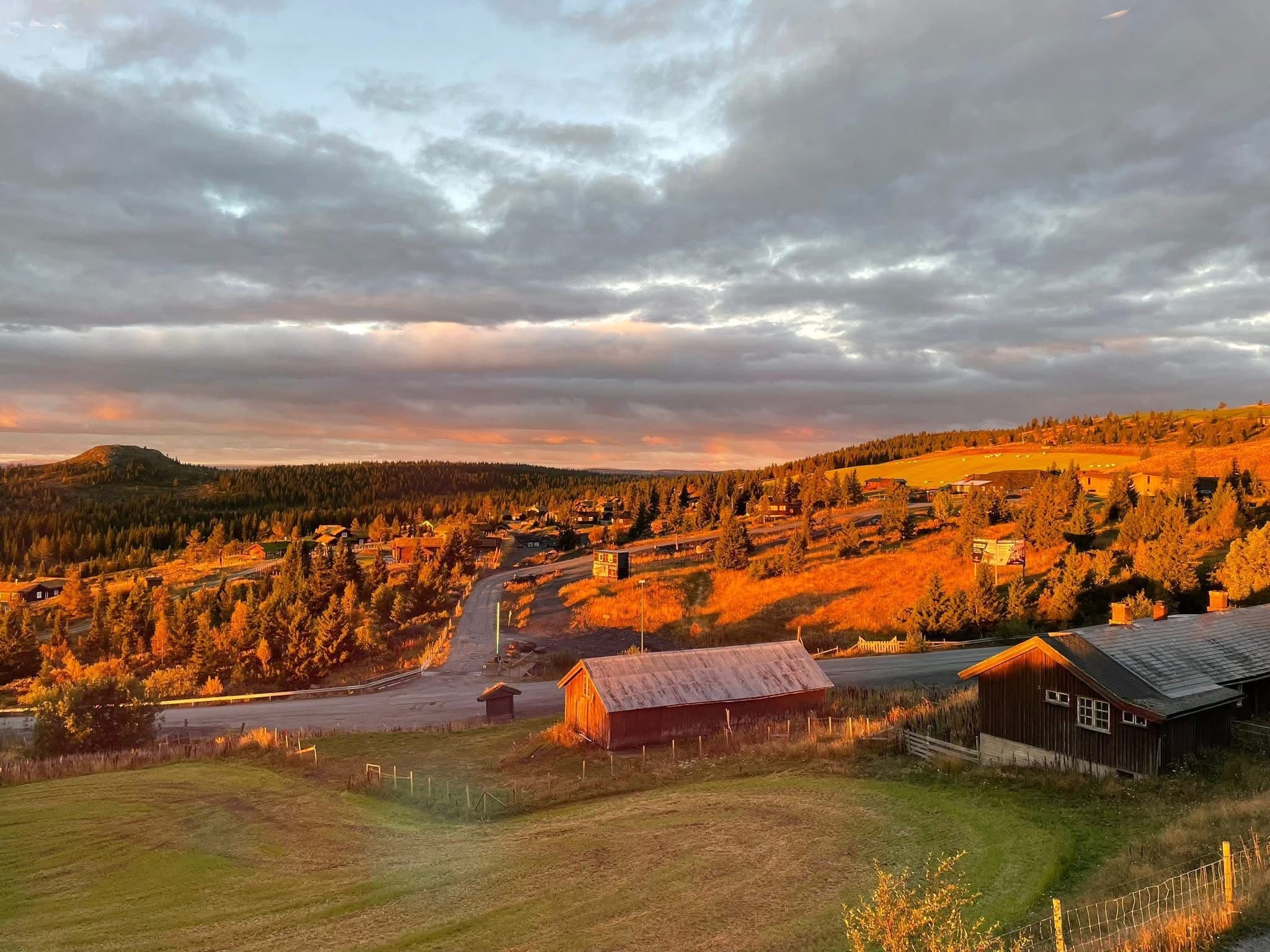 Sunrise lighting a rural valley with wooden barns, a winding road, and forested hills in Norway.