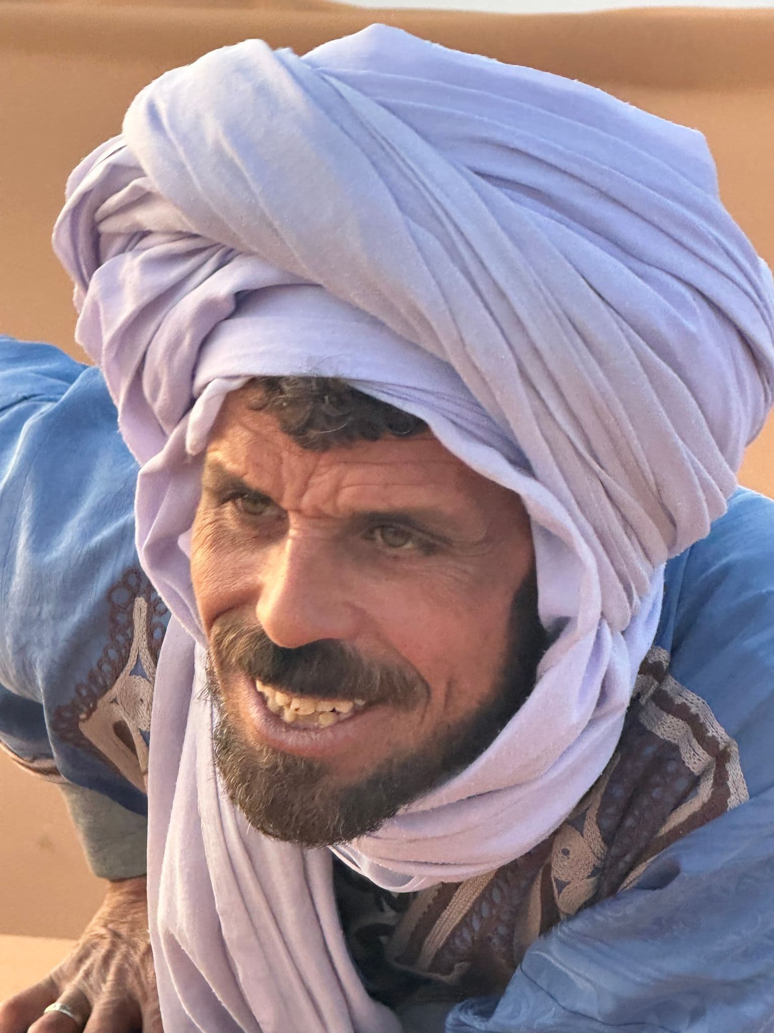 Man wearing a large light-purple turban leaning forward in the Sahara Desert, Morocco.