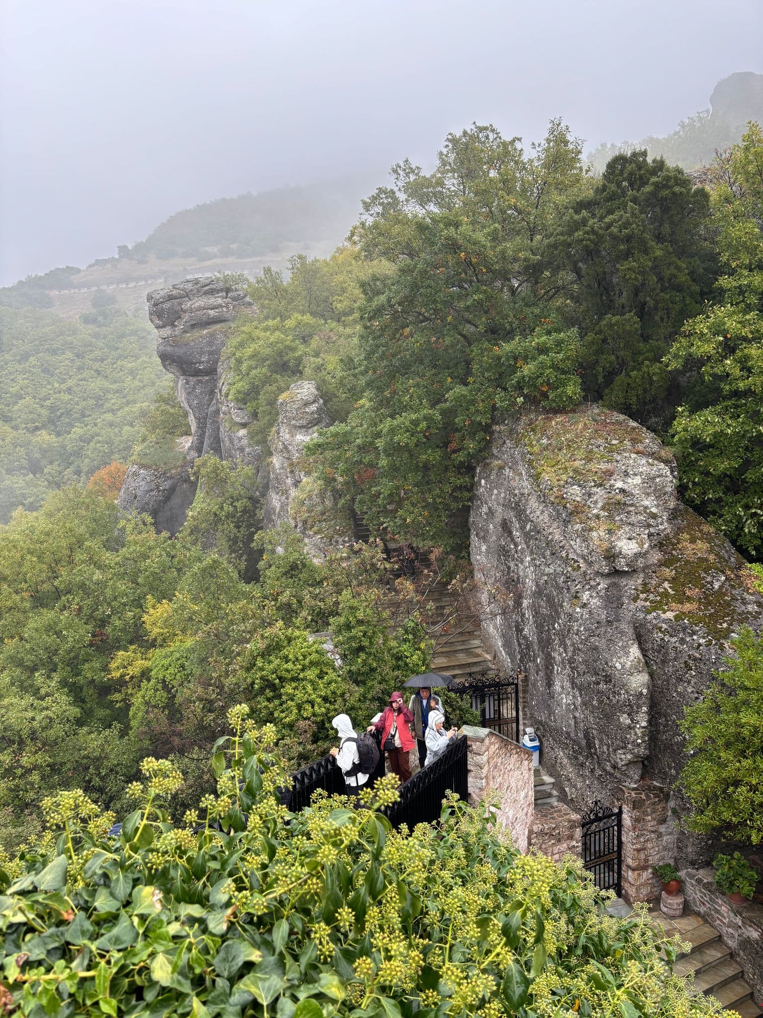 Monastery of Varlaam at Meteora with tourists on a stone terrace and steps amid misty cliffs near Kalambaka, Greece.