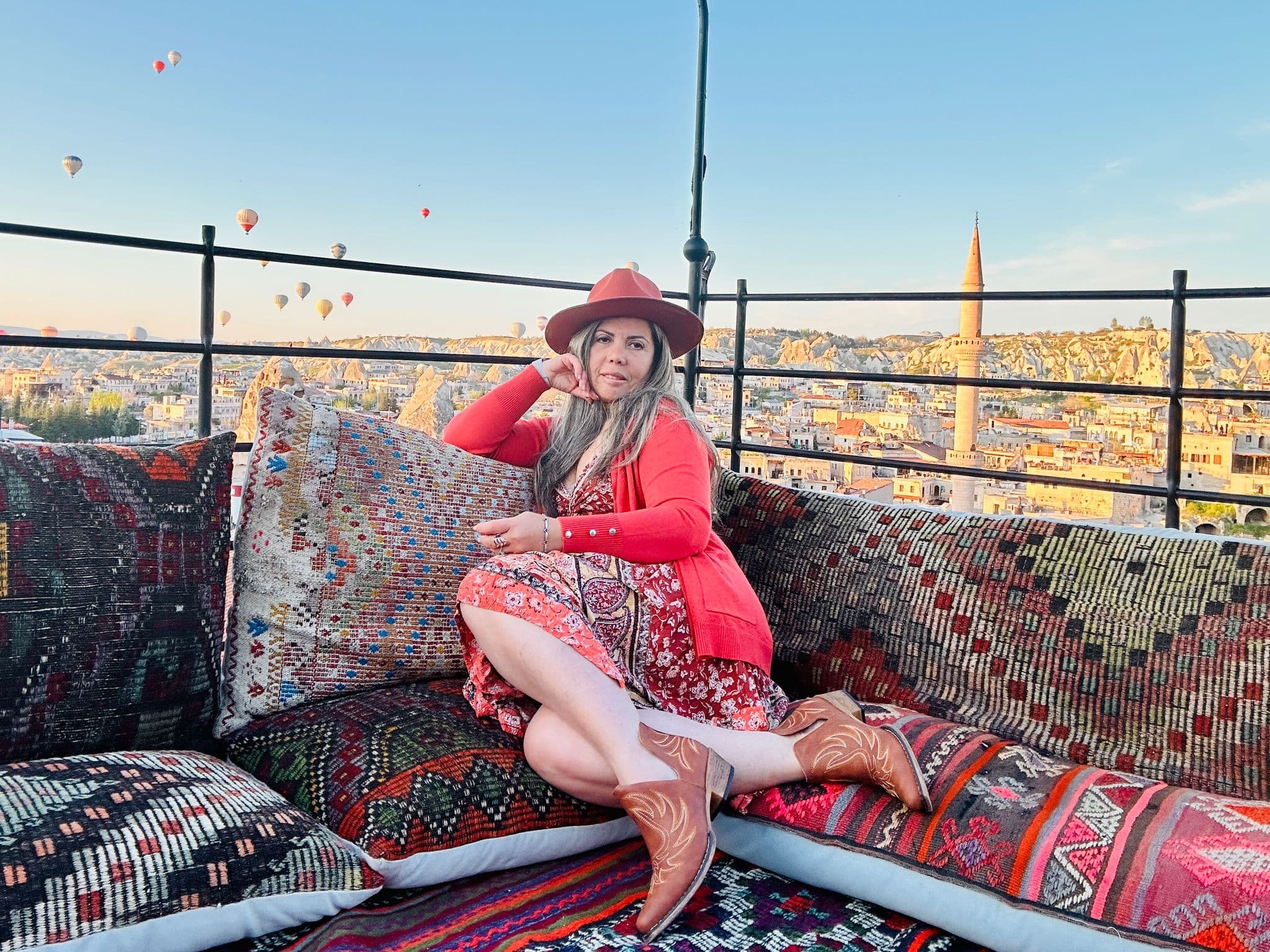 Hot air balloons over Cappadocia with a woman seated on a patterned rooftop terrace at sunrise in Göreme, Turkey.