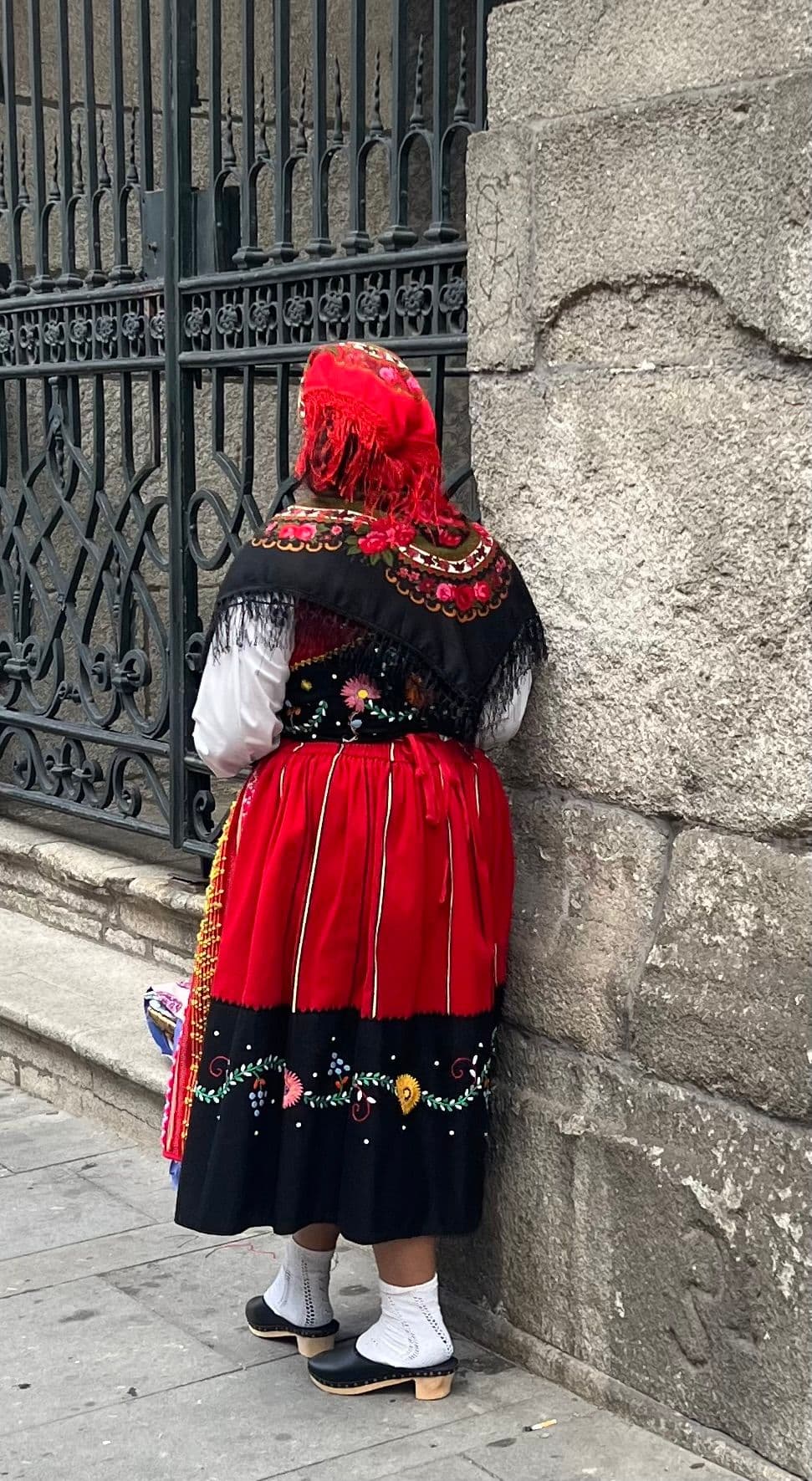 Woman in a red and black traditional Portuguese folk costume standing with her back to the camera by a stone wall and iron gate, Portugal.