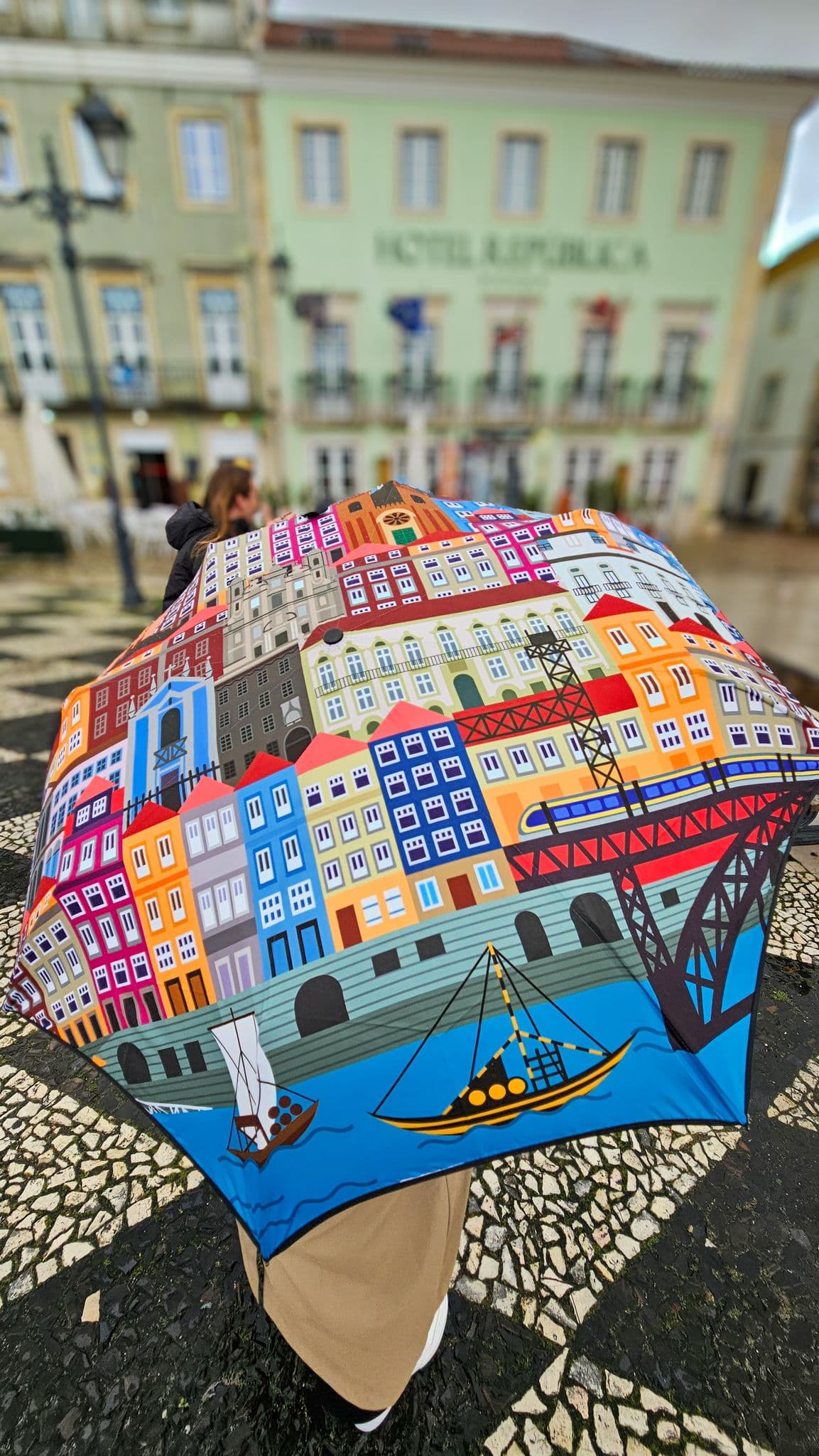 Colorful illustrated umbrella held over a person in Praça da República, Tomar, Portugal, with the Hotel República façade visible behind.