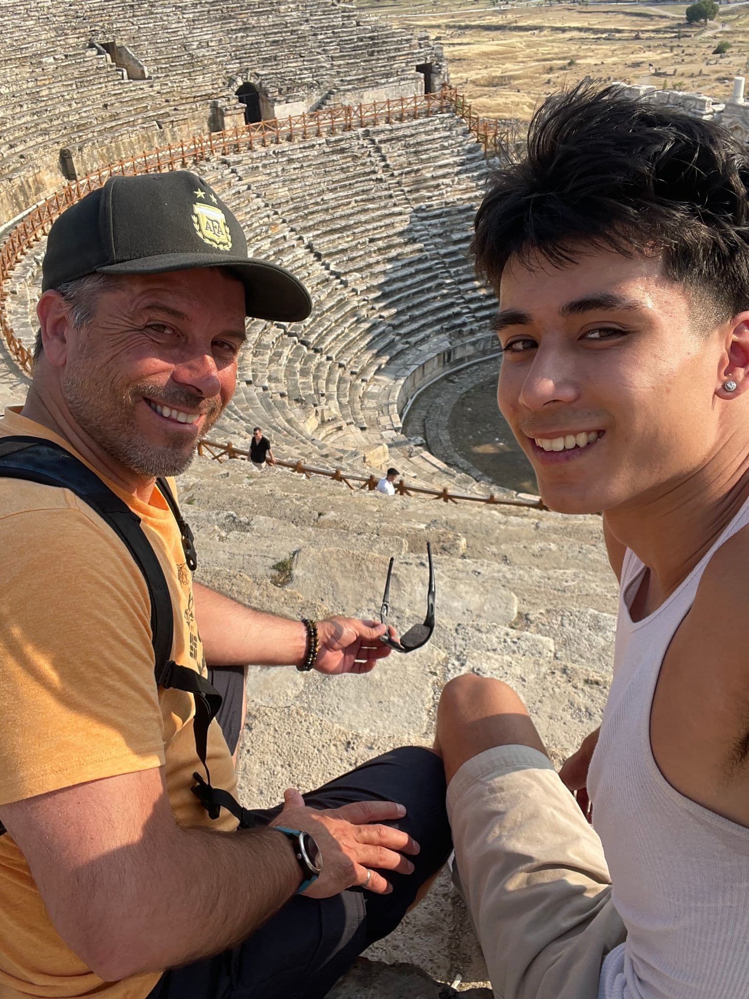 Ancient Roman amphitheater with two travelers sitting on stone tiers smiling for a selfie, historic site in Turkey.