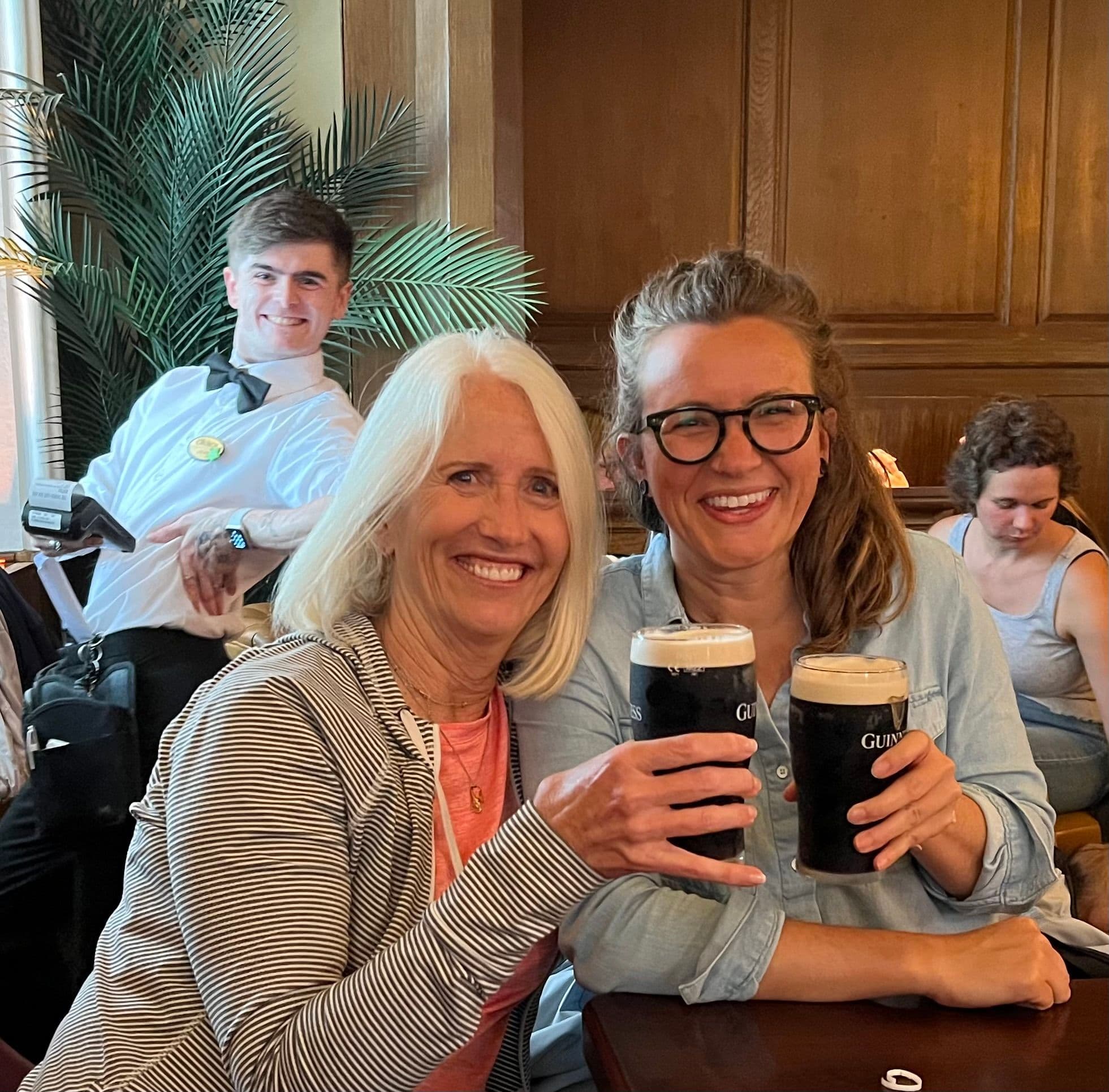 Two women clinking pints of Guinness in a wood-paneled Irish pub, a waiter photobombing behind them — Ireland.