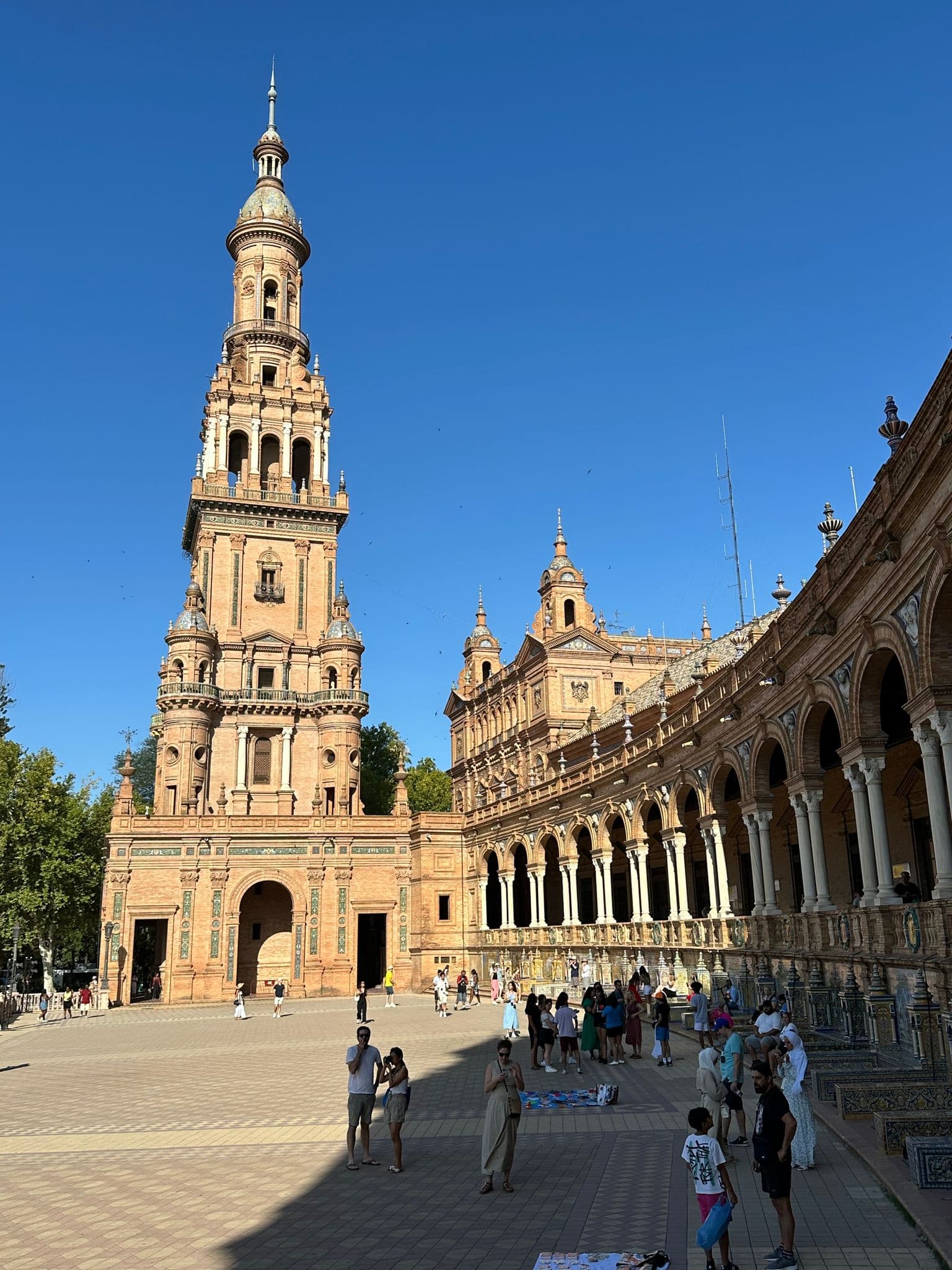 Plaza de España's tall tower and curved colonnade in Seville, Spain, with people walking and gathering in the sunny plaza.