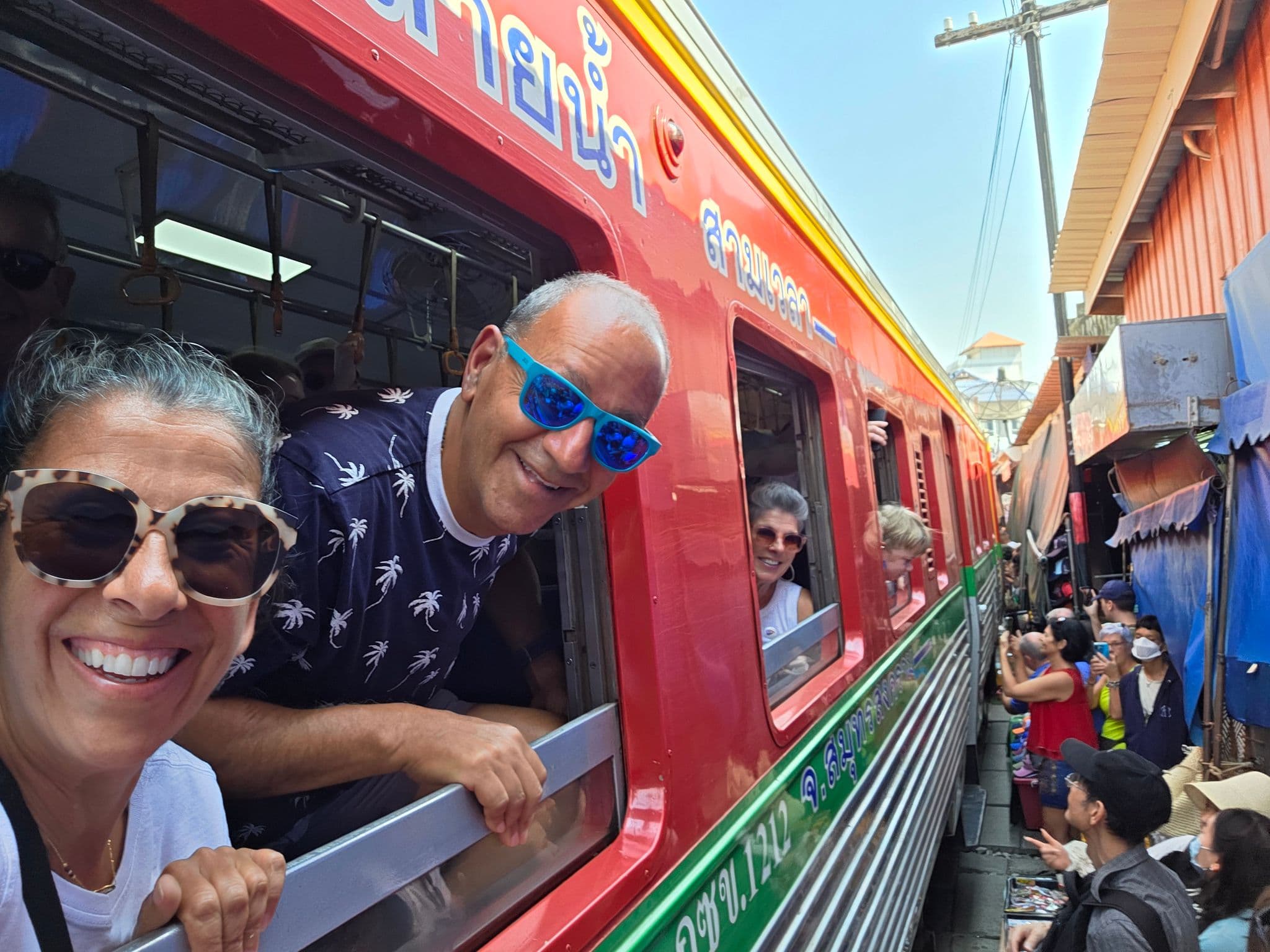 Red passenger train with travelers leaning from open windows as a crowded market squeezes alongside at Maeklong Railway Market, Thailand.