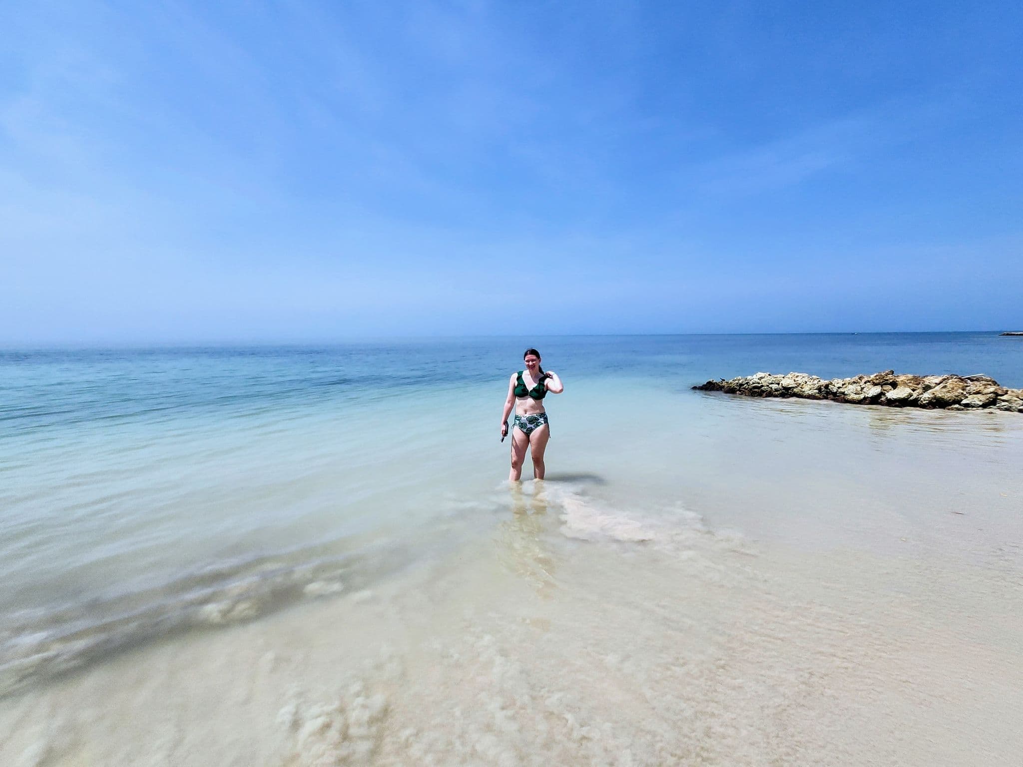 Woman standing in shallow clear water on a sandy beach near a rocky outcrop under a blue sky.