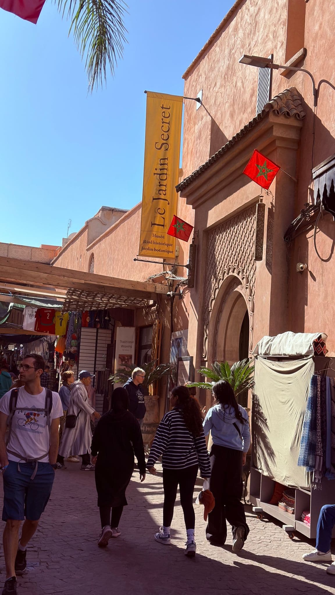 Le Jardin Secret entrance and banner with shoppers walking through a busy Medina street in Marrakech, Morocco.