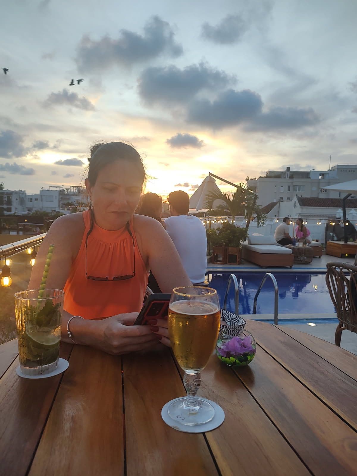 Woman at a rooftop table checking her phone beside drinks with a sunset over a pool in Cartagena, Colombia.