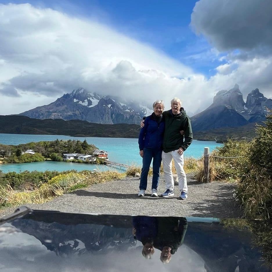 Turquoise lake and Torres del Paine peaks with two travelers standing arm-in-arm on a trail in Torres del Paine National Park, Chile.