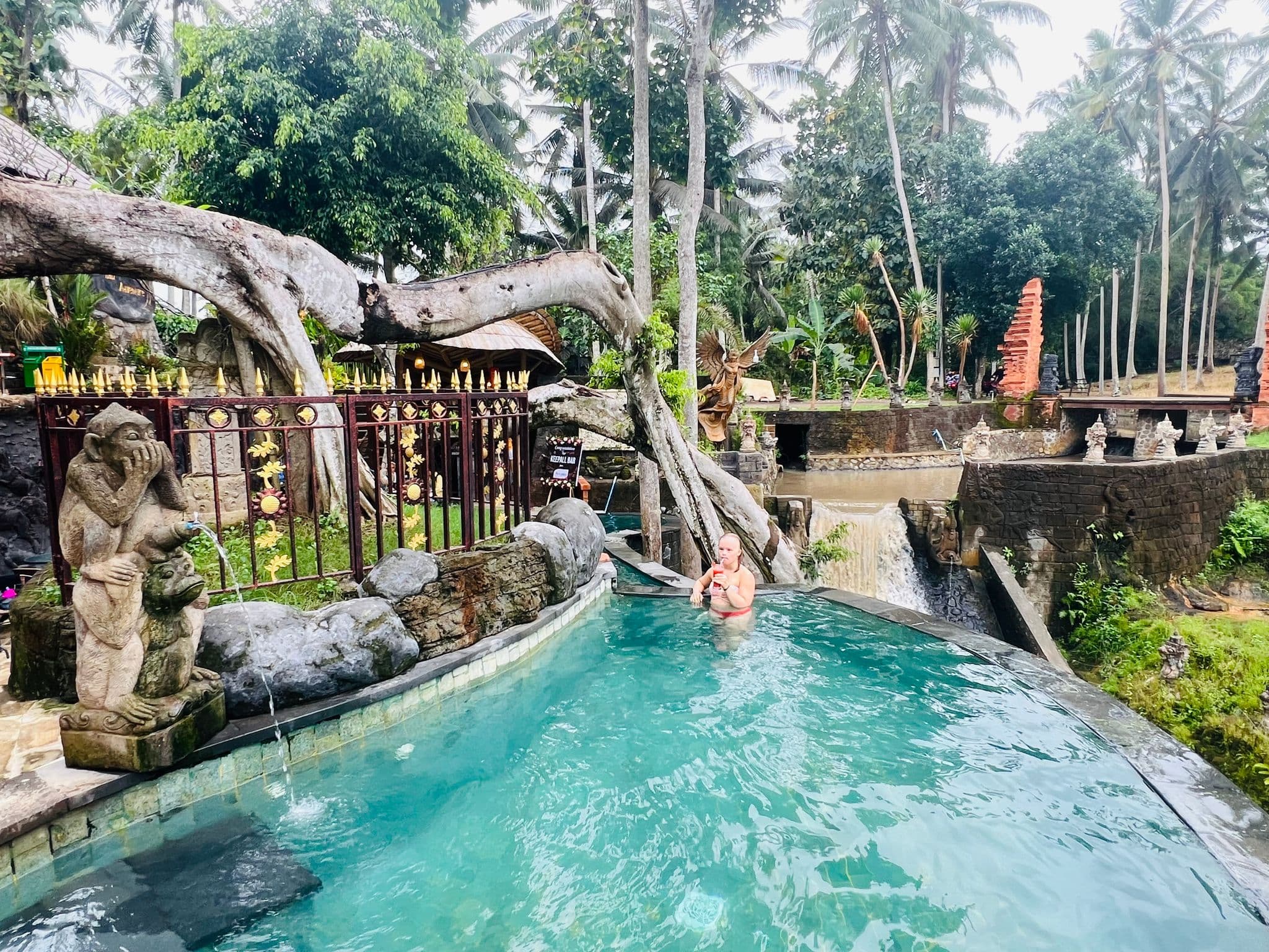 Infinity-style pool with a woman sipping a drink beside a small waterfall and carved Balinese statues in Bali, Indonesia.