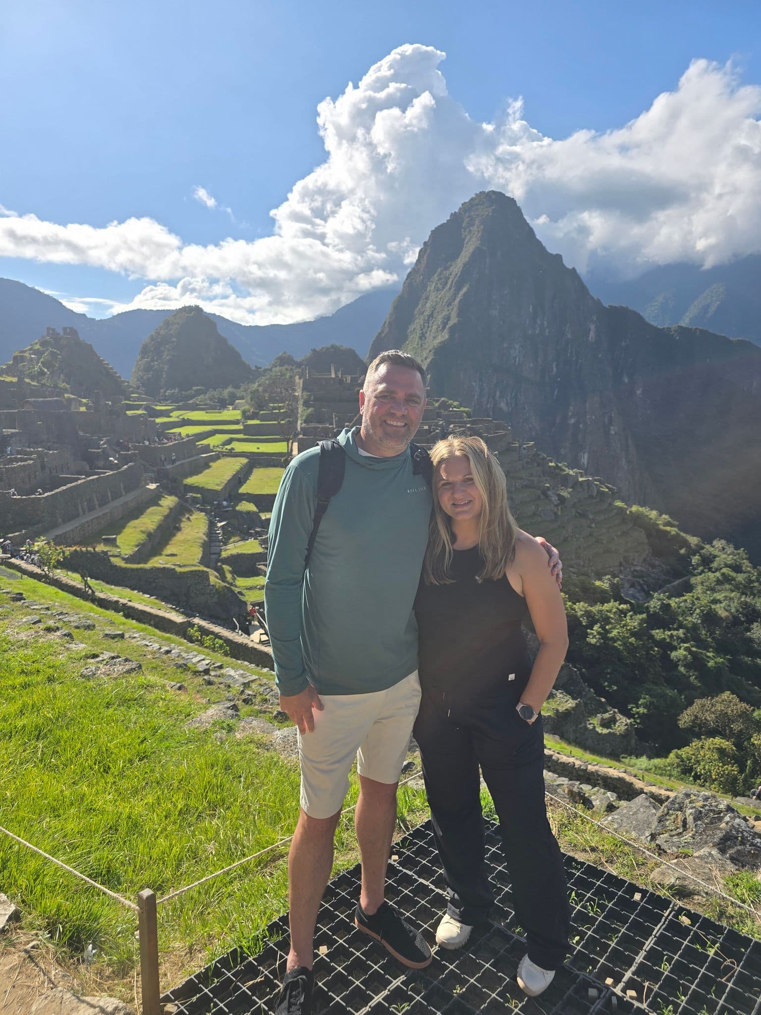 Machu Picchu ruins with Huayna Picchu behind and two travelers posing in the foreground, Cusco Region, Peru.