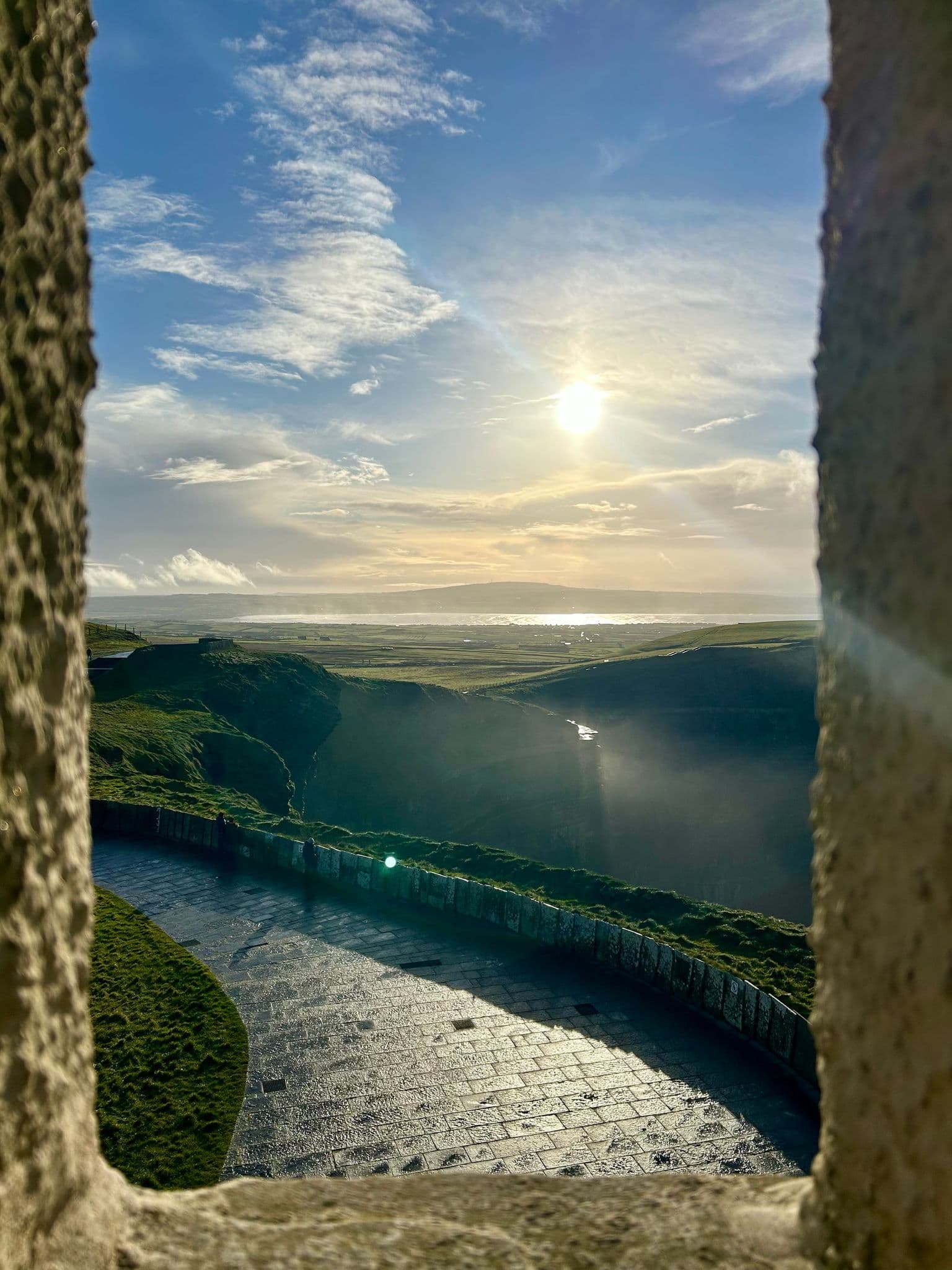 Cliffs of Moher seen through a stone window opening with sunlit sea and a curved cliff-top path in County Clare, Ireland.