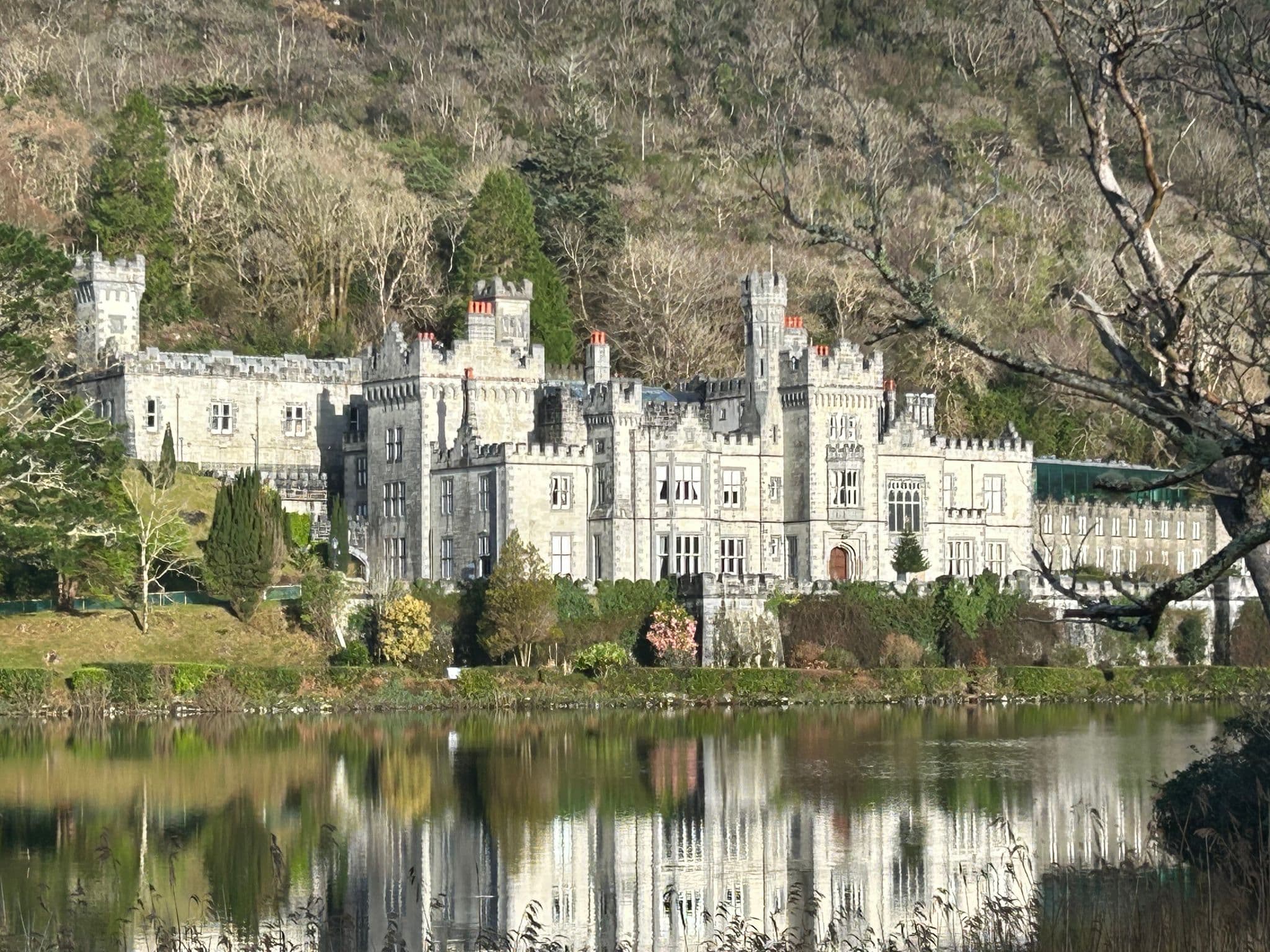 Large stone castle by a reflective lake with wooded hills behind, viewed on a trip in Ireland.