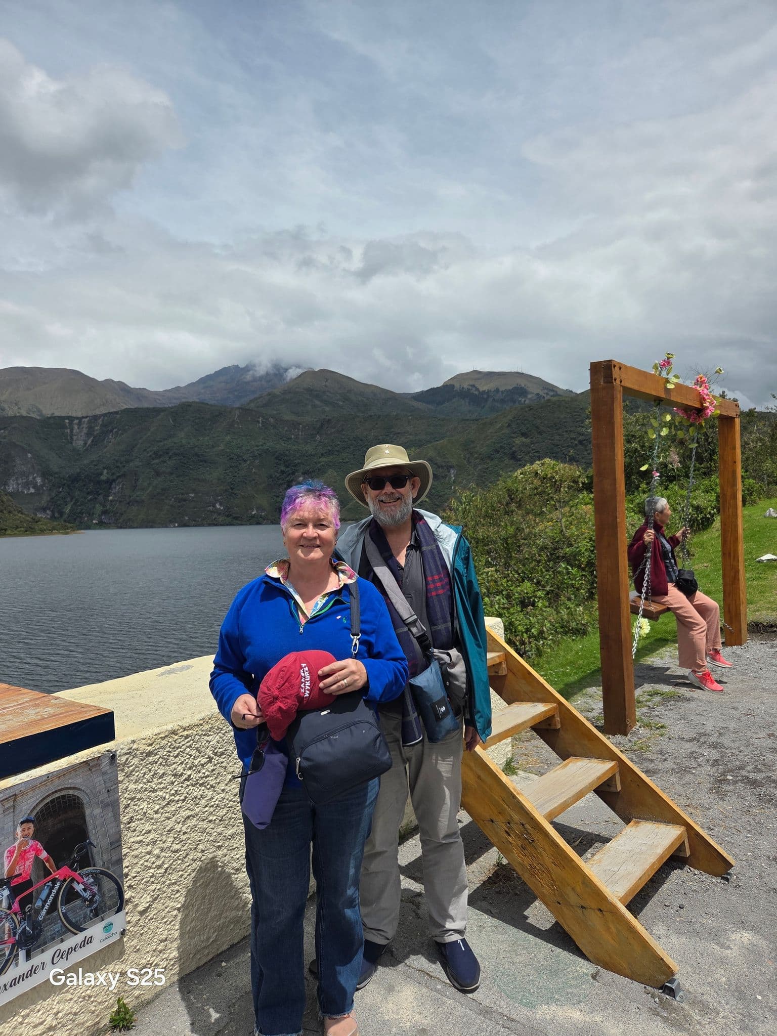 Two travelers standing by a mountain lake with green Andean hills behind them, wooden steps and a flowered swing at the side.