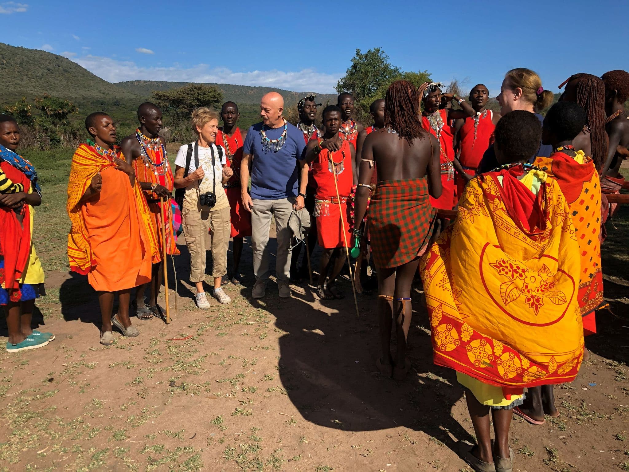 Maasai villagers in red shukas and two tourists standing and talking on a grassy plain near rolling hills, Kenya.