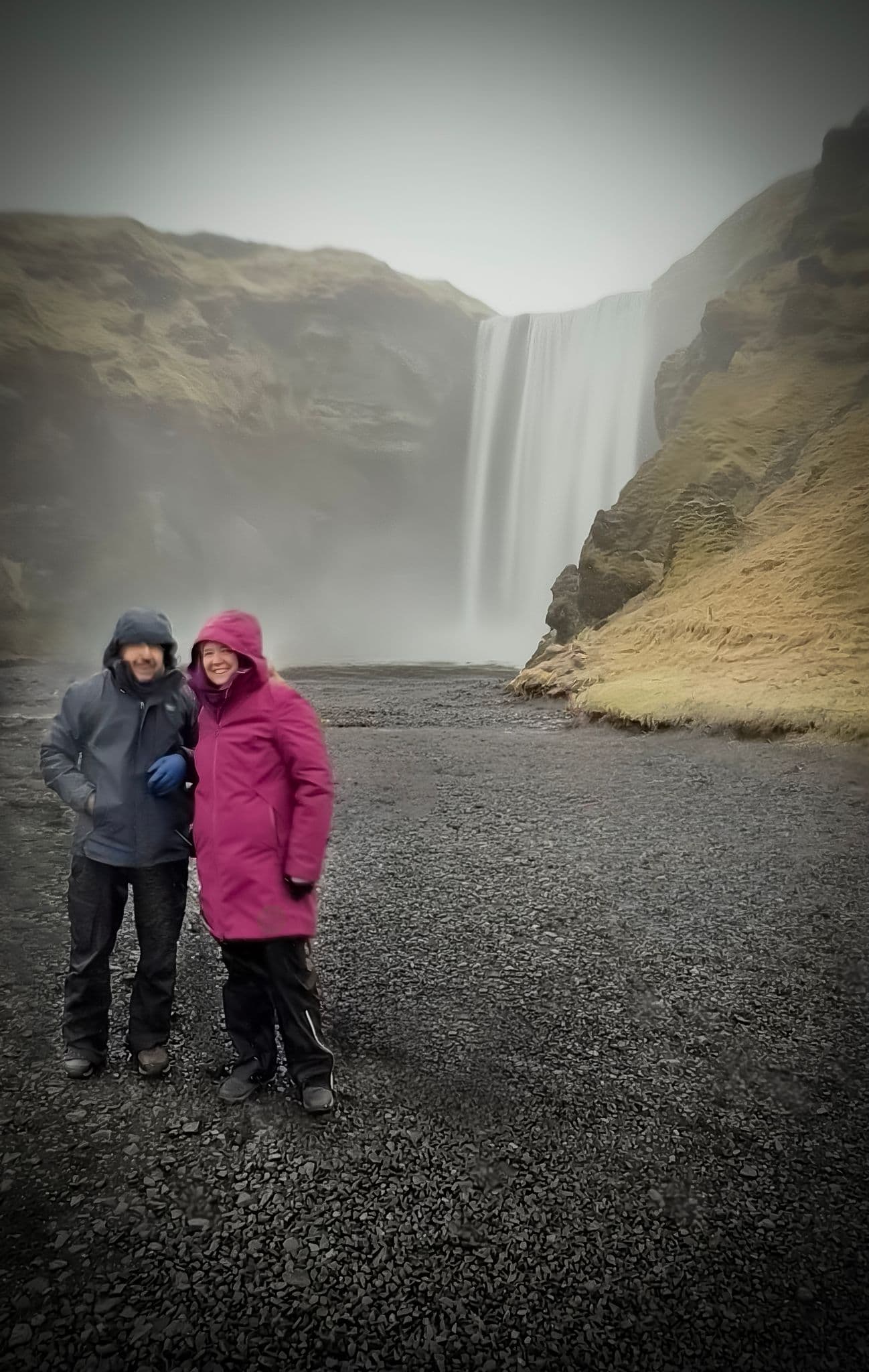 Skógafoss waterfall with two bundled travelers standing on black gravel near the falls on a windy day, Skógafoss, Iceland.